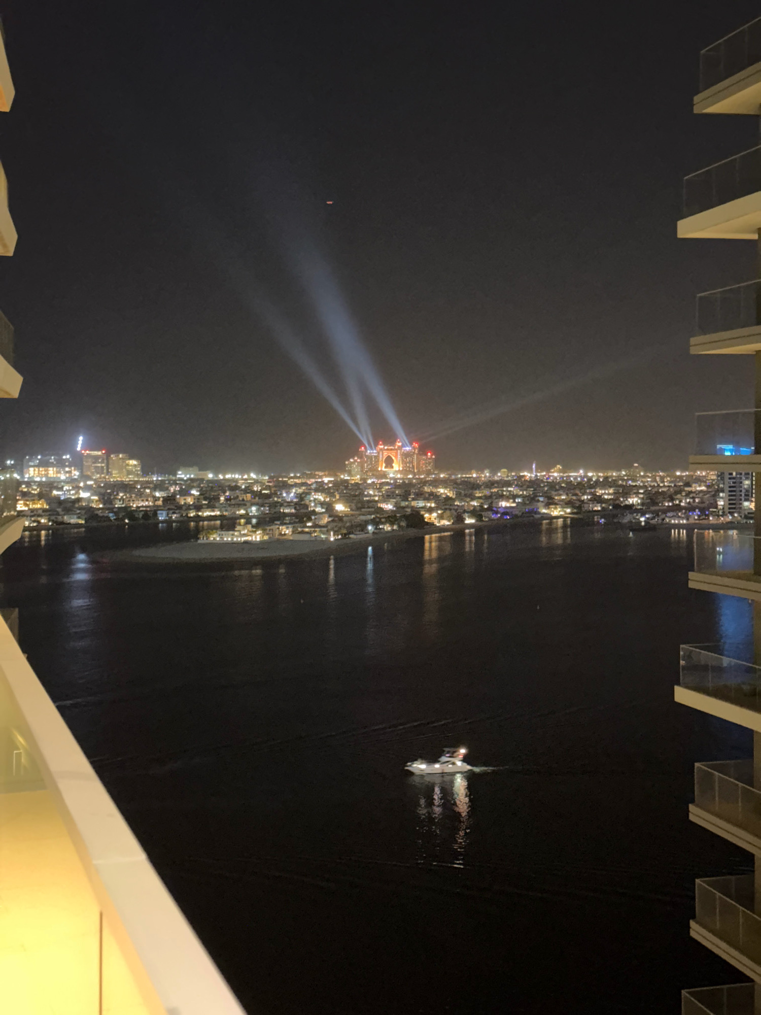 A nighttime aerial view captures a cityscape in Dubai, United Arab Emirates, illuminated by numerous lights. In the foreground, the balconies of a modern building create a framing effect on the left and right sides. A body of dark water dominates the lower half of the image, with a small, illuminated boat navigating its surface, leaving a subtle wake.
Across the water, the sprawling city unfolds. The distant skyline is dotted with buildings of varying heights, their windows and external lights creating a warm glow against the night sky. Prominent in the center-right background are two distinct structures featuring dramatic light beams projecting upwards into the darkness, suggesting a special event or landmark illumination. The overall impression is one of a vibrant and active urban environment at night, with the water serving as a reflective element and a path for marine activity.