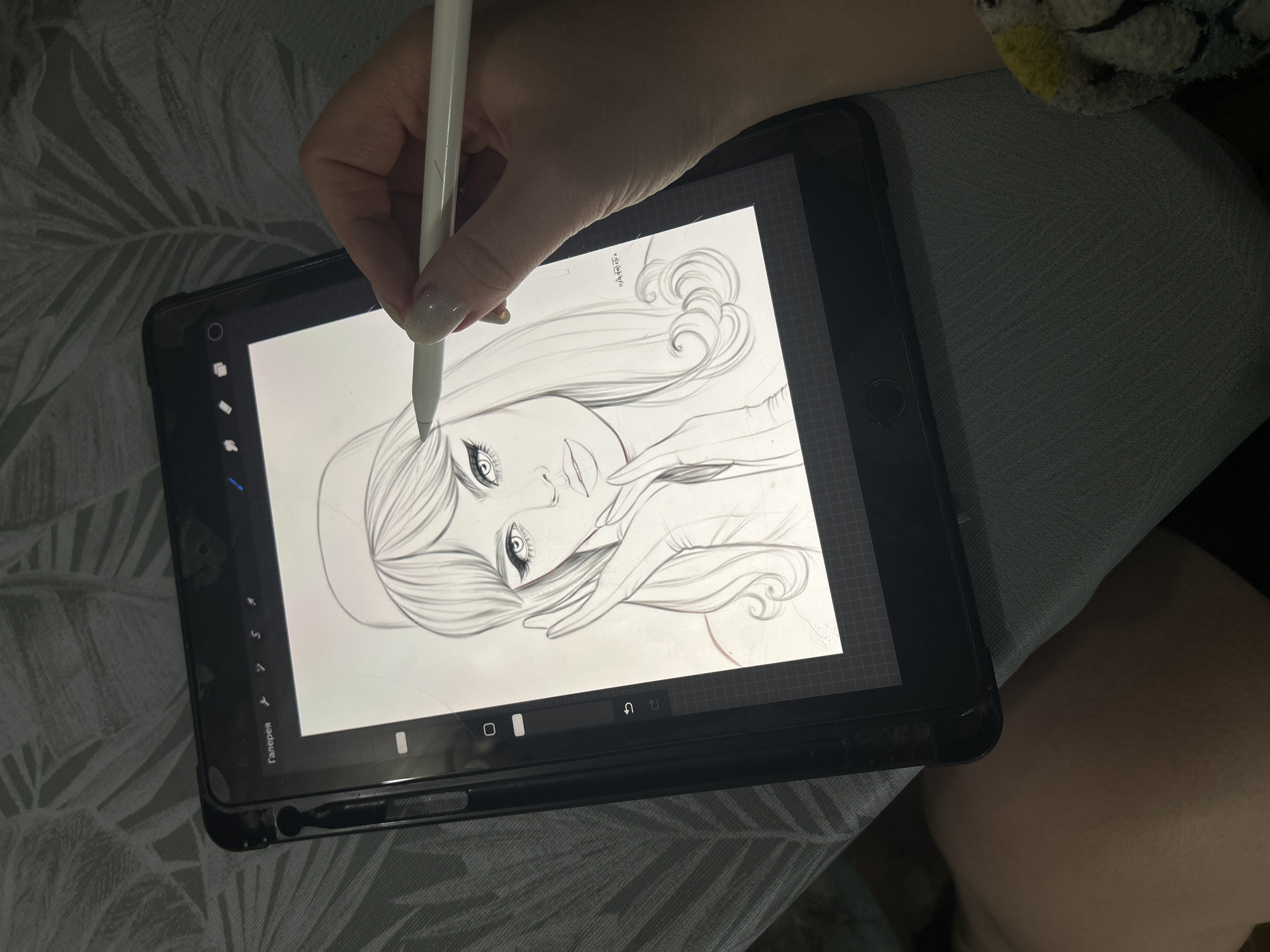 A person is drawing a sketch of a woman's face on a black tablet with a white stylus. The tablet screen displays the drawing, which shows a woman with detailed eyes and flowing hair. The tablet is in a black protective case and rests on a surface with a leaf-like pattern. The person's hand holds the stylus, positioned to draw on the tablet. The environment is indoors, and the lighting is soft. There's no discernible information to identify the location as the city of Фоки, Russia.