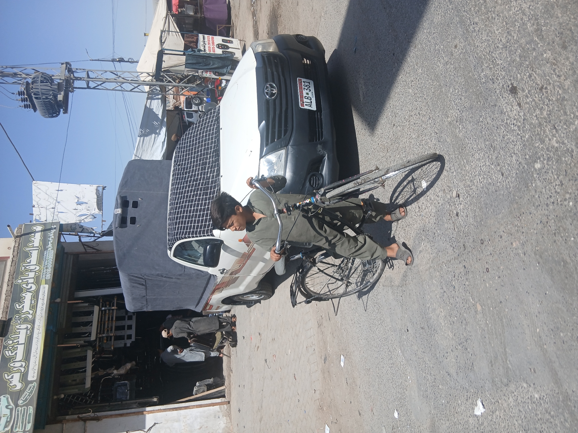 A street scene in Quetta City Tehsil, Pakistan, presents a juxtaposition of elements. A young man wearing an olive-green outfit is seated on a bicycle. A white and black pickup truck with the license plate "ALB-381 Sindh" is parked behind him, partially obscured by a dark covering or equipment mounted on its bed. To the left, a shopfront features Urdu script signage, indicating a business establishment. Overhead, power lines and a transformer dominate the sky. The street is paved, showing signs of wear and tear. The day is sunny, casting shadows on the ground.