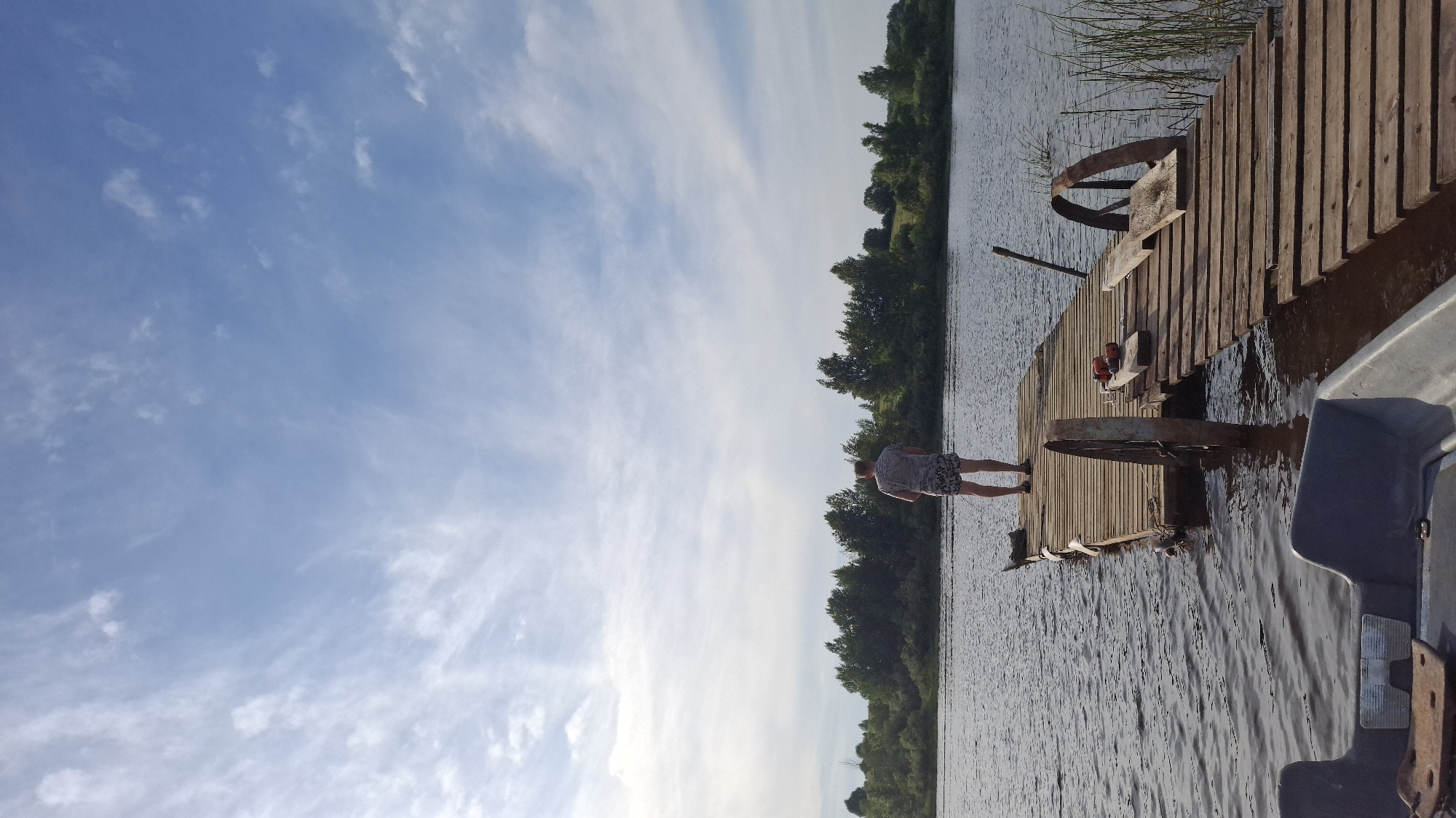 A person stands on a wooden pier extending into a calm body of water, facing away from the camera. The pier is weathered and appears to be constructed from planks. Two large, rusty metal wheels are attached to the pier's structure. In the foreground, the bow of a grey boat is visible. The background features a dense line of green trees and vegetation along the opposite bank of the water. The sky above is a light blue with scattered white, wispy clouds. The scene appears to be an outdoor setting in a natural environment, likely during daylight hours. The location is identified as Sitno, Russia.