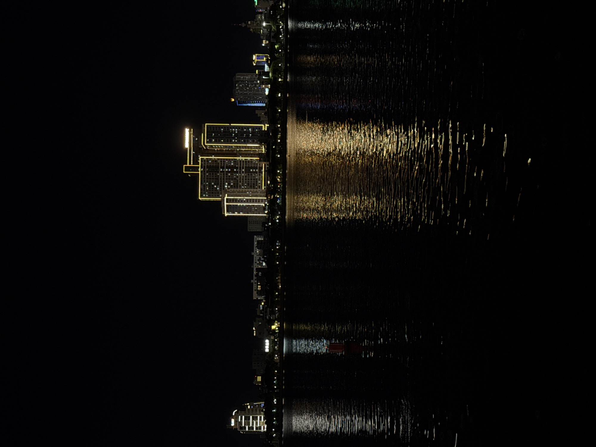 The image captures a nighttime cityscape with a body of water in the foreground. The water reflects the lights of the buildings, creating shimmering patterns. In the background, a cluster of modern high-rise buildings are illuminated with yellow and blue lights. Some of these buildings are outlined with bright yellow lights, highlighting their architectural features. The sky is dark, indicating it is nighttime. A red buoy with a white band is visible in the water, slightly to the left of the center. The scene appears to be a waterfront area in the city of Đà Nẵng, Vietnam, likely along the Han River, given the presence of modern illuminated skyscrapers and the general urban waterfront aesthetic.