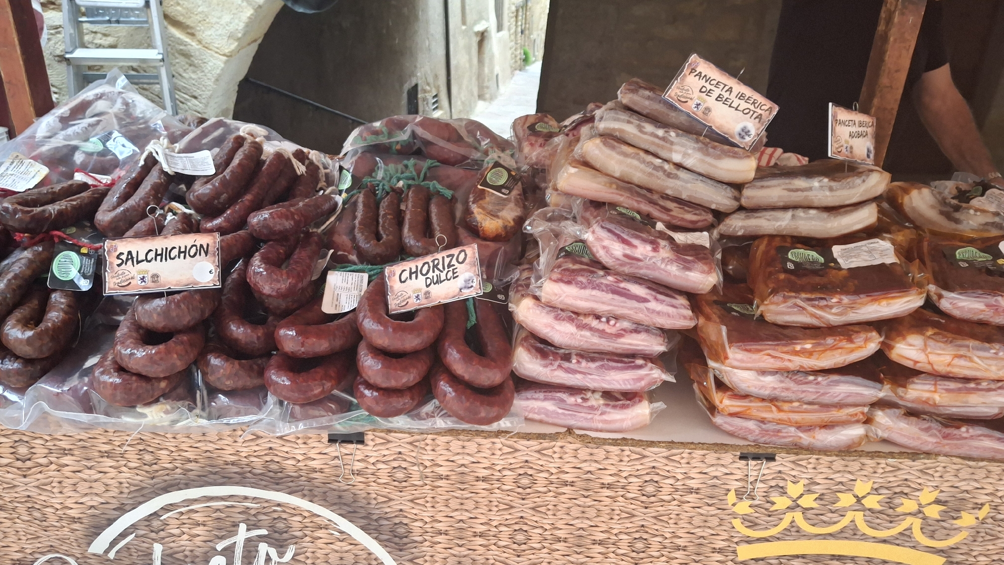 The image depicts a market stall displaying a variety of cured meats, likely in the city of La Fresneda, Spain, given the Spanish labeling. The foreground shows a wicker-covered counter with several clips holding up signage and packaging. Prominently displayed are rows of sausages. On the left, several strings of dark red sausages are visible, some with small labels. A larger sign reads "SALCHICHÓN". To the right of the salchichón, a sign reads "CHORIZO DULCE". Behind the sausages, there are stacked portions of cured pork belly and other cuts of meat, individually packaged. Some of these packages have labels with Spanish text, including "PANCETA IBÉRICA DE BELLOTA" and "PANCETA IBÉRICA ADOBADA". The background suggests an outdoor market setting, with a stone archway and buildings visible, characteristic of historic towns in Spain. The lighting appears to be natural daylight. No people are clearly visible, though a person's arm is partially seen on the far right, suggesting a vendor.