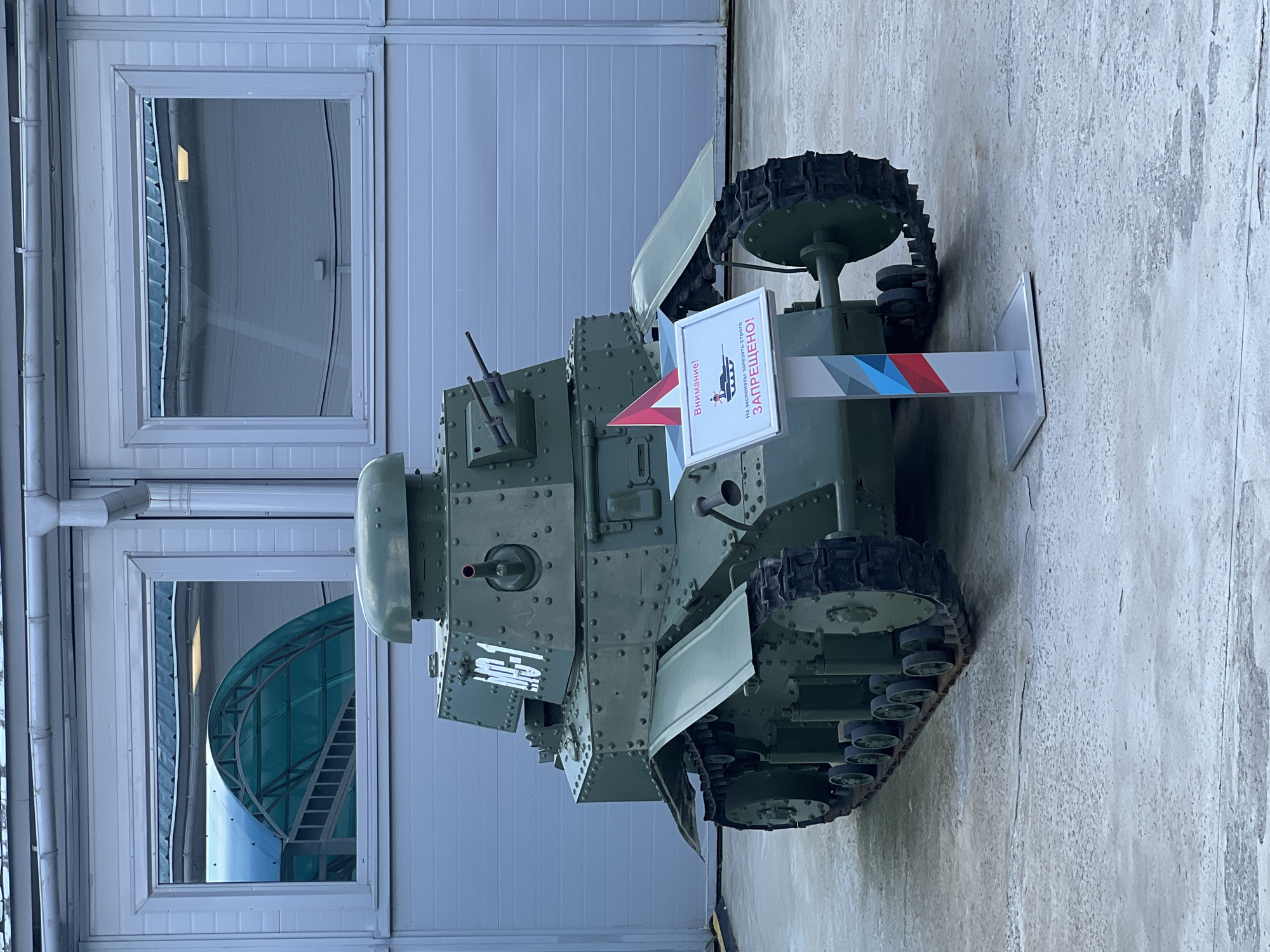 An olive-drab Soviet-era light tank, identified as a T-18 (MS-1) by the markings on its turret, is positioned outdoors on a paved surface in front of a modern building with large, reflective windows. The tank, which appears to be a museum exhibit, has visible rivets along its hull and turret, and two machine guns mounted on the front of the turret. A white sign with red and blue triangular accents stands in front of the tank, featuring a drawing of a person standing on the tank and Russian text that translates to "Attention! Climbing on exhibits is FORBIDDEN!". The background shows the exterior of a building with a corrugated metal facade and two large windows reflecting an overhead structure. The location is specified as the Patriot Exhibition Center in Kubinka, Russia. There are no people in the image.