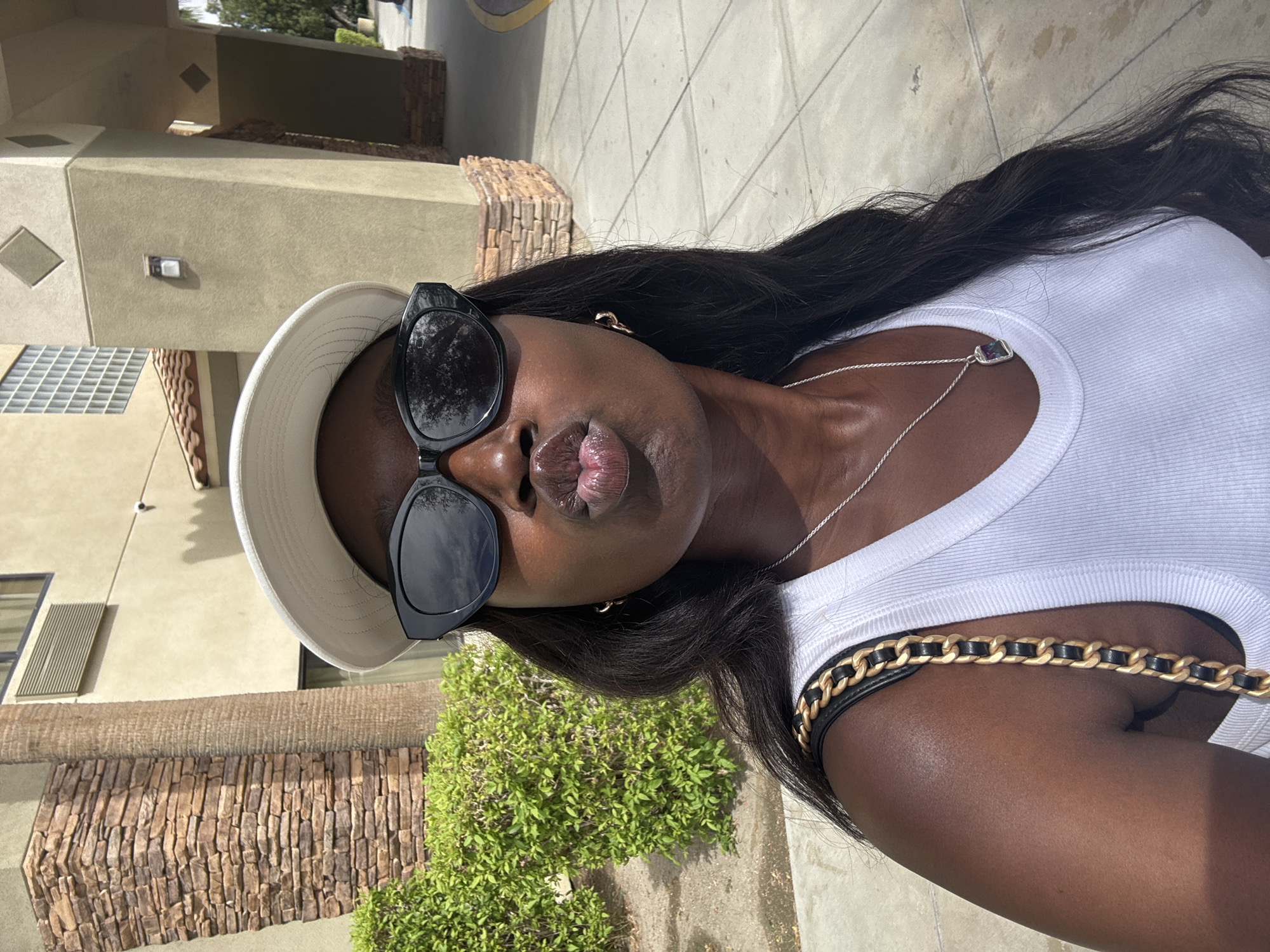 A woman with dark skin, dark hair, and wearing a white tank top, a white bucket hat, and sunglasses stands on a sidewalk in what appears to be Palm Desert, California. She is facing the camera with her lips pursed. She has a silver necklace with a stone pendant and a black purse with a gold chain strap. The building behind her is light brown with brick accents. A small green bush is in the foreground to the left of the frame, and a sidewalk is visible in the upper right.