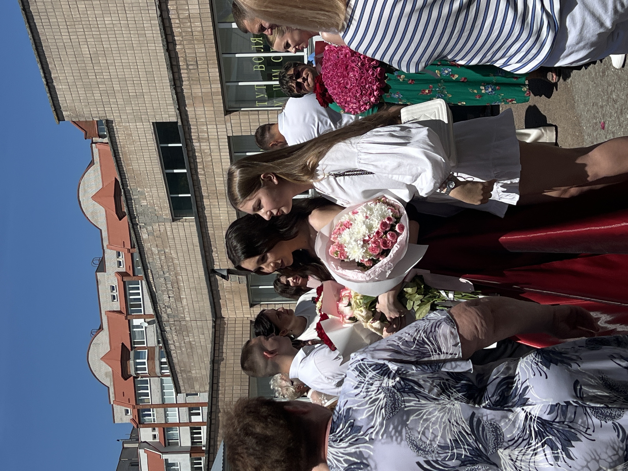 This image captures a group of people gathered outdoors, possibly for a celebratory occasion. Several individuals are holding bouquets of flowers, with one prominent bouquet featuring pink and white roses and white chrysanthemums. The people are dressed in casual attire, including a white dress and a striped shirt. Some individuals appear to be young women, possibly students.
The scene is set against a backdrop of a light-colored brick building with a distinctive reddish-brown architectural detail at the top, suggesting an urban or semi-urban environment. The clear blue sky indicates it is daytime, and the strong shadows suggest bright sunlight, implying fair weather.
One individual in the background wears sunglasses and a white t-shirt. Another person is wearing a green dress with a floral pattern. The red surface in the foreground might be part of a car or bench.
Visible text on the building includes Cyrillic letters, with "ТУТ ВОЛЯ" partially legible. This text, combined with the mention of Ternopil, Ukraine as location context, suggests the event may be taking place in Ukraine. The overall mood appears to be happy and festive, as suggested by the flowers and the gathering of people.