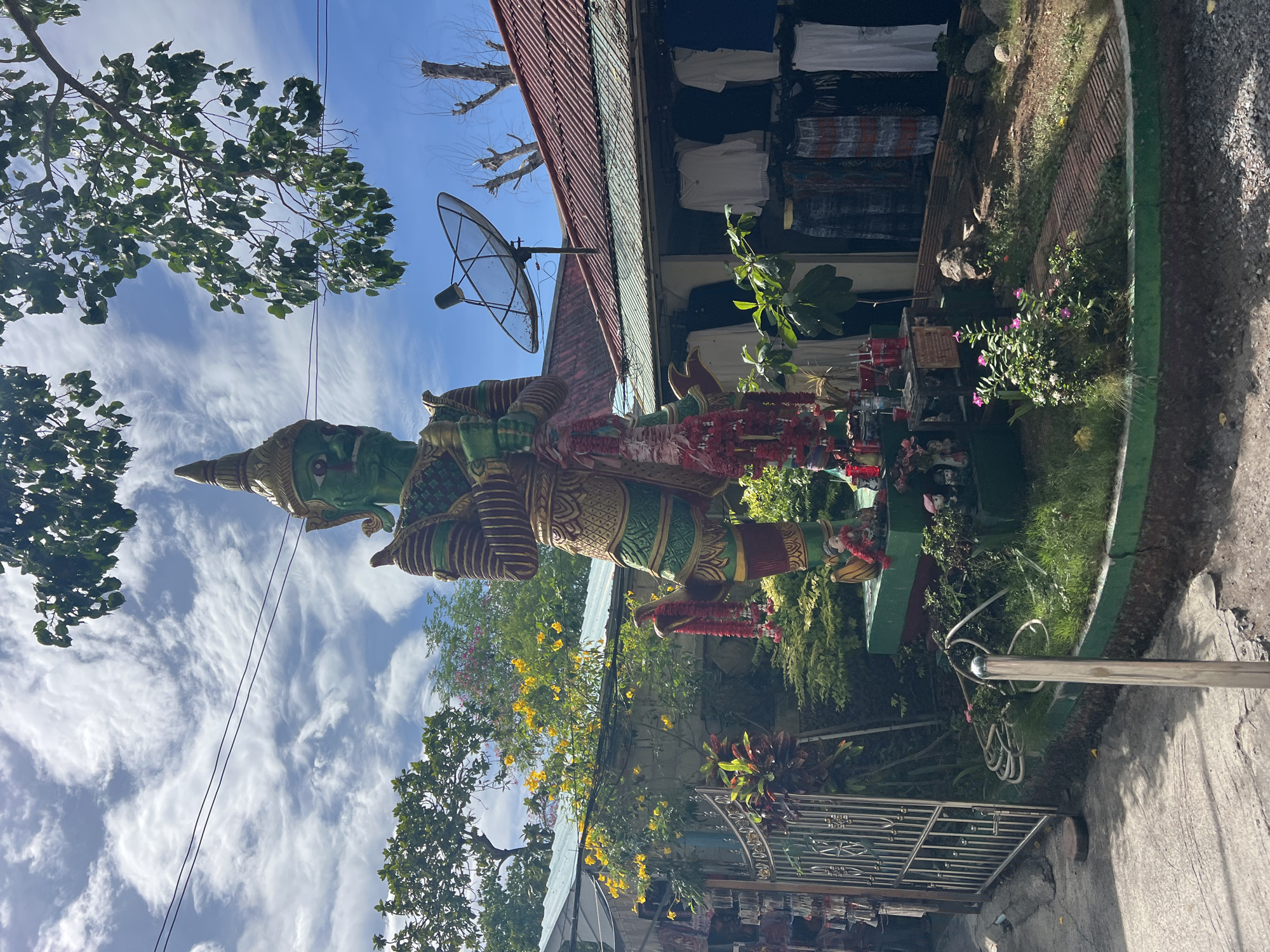 This is a full-shot image of a large, green, statue of a guardian deity standing outside a row of shops in Baan Plai Laem, Thailand. The statue is ornately decorated with gold and red accents and is adorned with garlands of red flowers. It is standing on a raised platform surrounded by greenery. Behind the statue, a satellite dish is mounted on the roof of a building. The shops appear to be selling clothing. The sky is blue with scattered white clouds, and trees are visible in the background. A metal gate is to the left of the statue. The scene appears to be outdoors on a sunny day.
