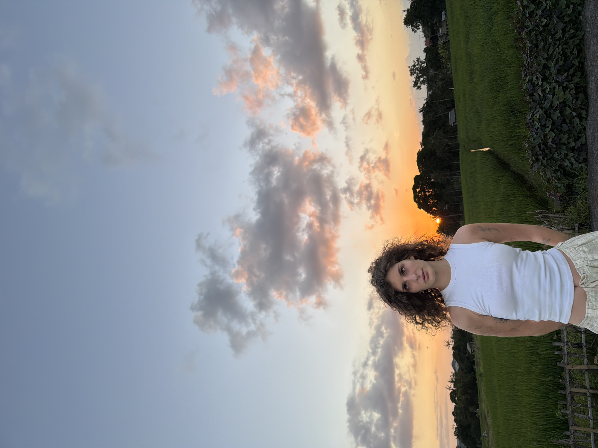 A medium shot captures a young woman with curly brown hair standing in a lush green rice field at sunset in North Kuta, Indonesia. She is wearing a white tank top and light-colored pants. The sky is filled with soft pink and orange clouds, and the sun is low on the horizon, casting a warm glow over the landscape. In the background, trees and buildings are silhouetted against the colorful sky. A wooden fence runs along the edge of the rice field. The overall atmosphere is serene and natural.