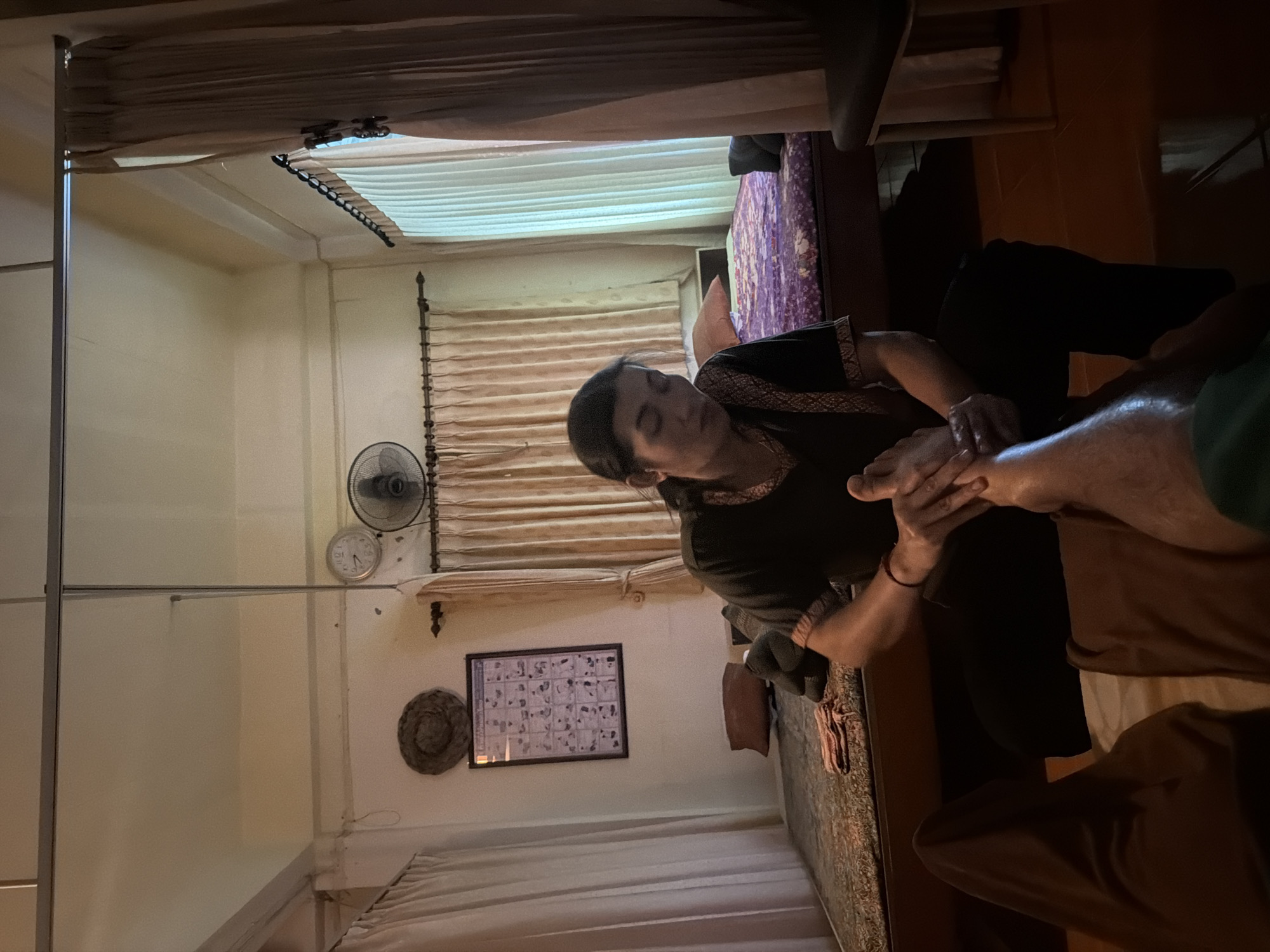 The image shows a foot massage being performed in a room in Baan Tai, Thailand. The room appears to be a massage studio, with two beds covered in patterned fabric and curtains hanging on the walls. A framed poster with a diagram of massage points is displayed on the wall. A person, likely a masseuse, is seated and giving a foot massage to another person whose legs are visible in the foreground. The lighting in the room is dim and warm. A ceiling fan is mounted on the wall.