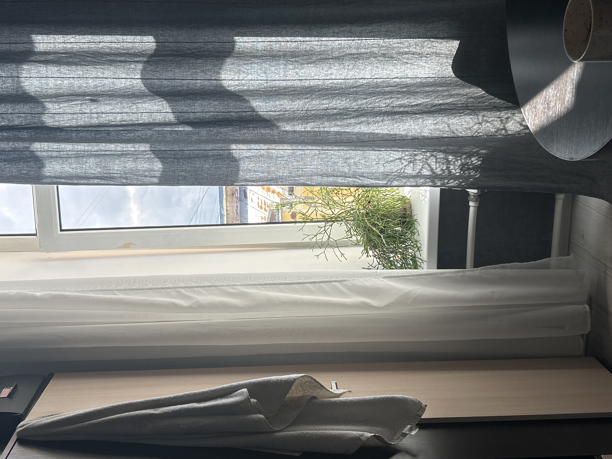 The image depicts an interior scene in Saint Petersburg, Russia, focusing on a window. The window is framed by two layers of curtains: a gray, textured blind partially drawn at the top and a sheer white curtain below. Outside the window, a view of buildings and foliage is visible, suggesting an urban setting. A green plant sits on the windowsill. In the foreground, a light gray folded towel rests on a light brown surface, likely a table or shelf. The overall lighting is soft, with shadows cast by the curtains and the sun outside. A round table is visible to the right with an object on it, possibly a cup.