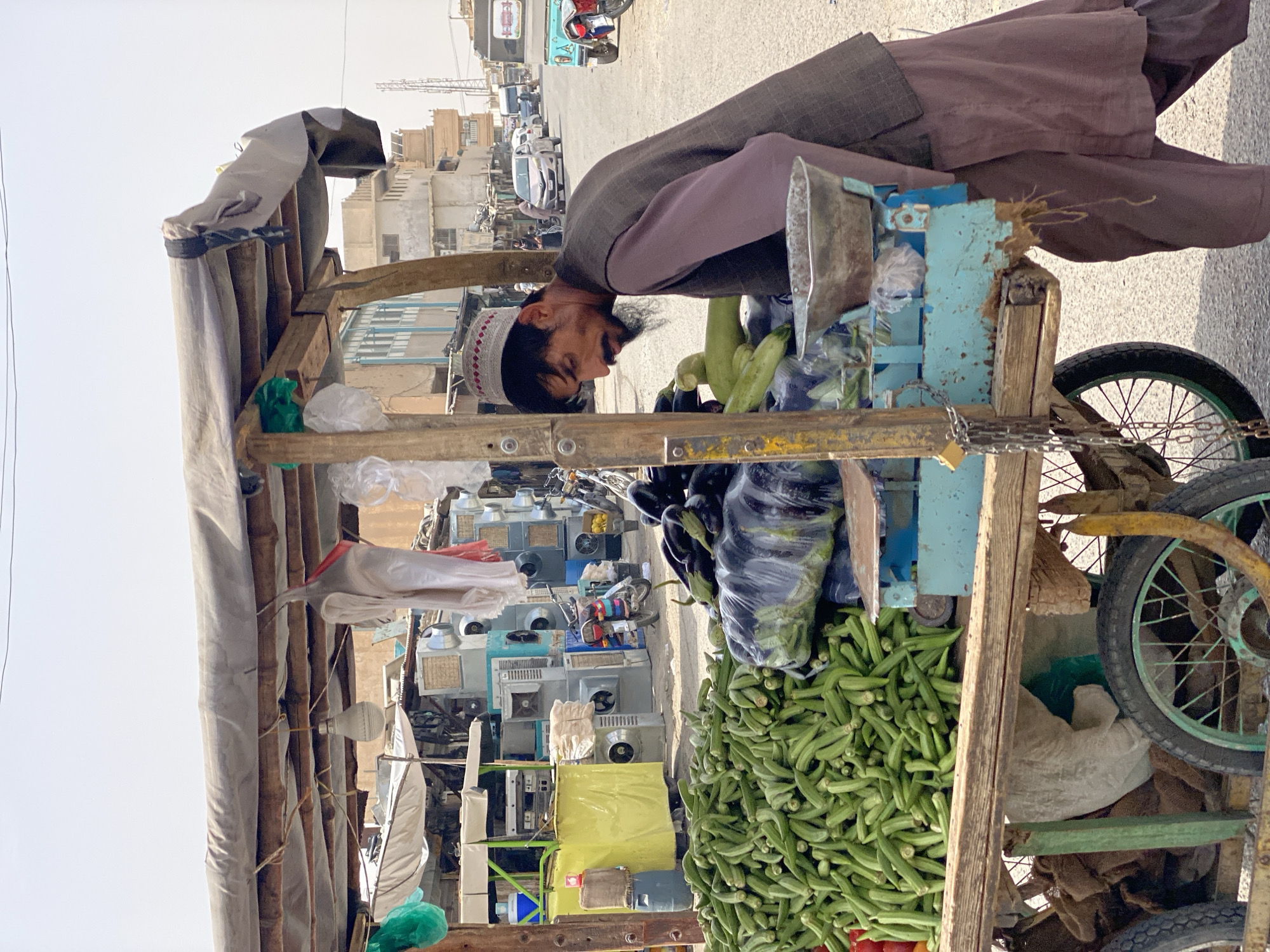 The image depicts a street scene in Kandahar, Afghanistan, featuring a man standing next to a vegetable cart. The cart is laden with a large pile of green okra in the foreground, with darker eggplants and green gourds visible behind it. A metal scale sits on the cart's blue platform. The man, wearing a brown vest over a mauve tunic and trousers, and a white and red patterned skullcap, is looking down at the vegetables. He has a mustache and a beard. The cart is made of wood and metal, with large wheels. In the background, a street is visible with buildings, parked motorcycles, and vehicles, suggesting a bustling market area. The sky is bright and overcast.