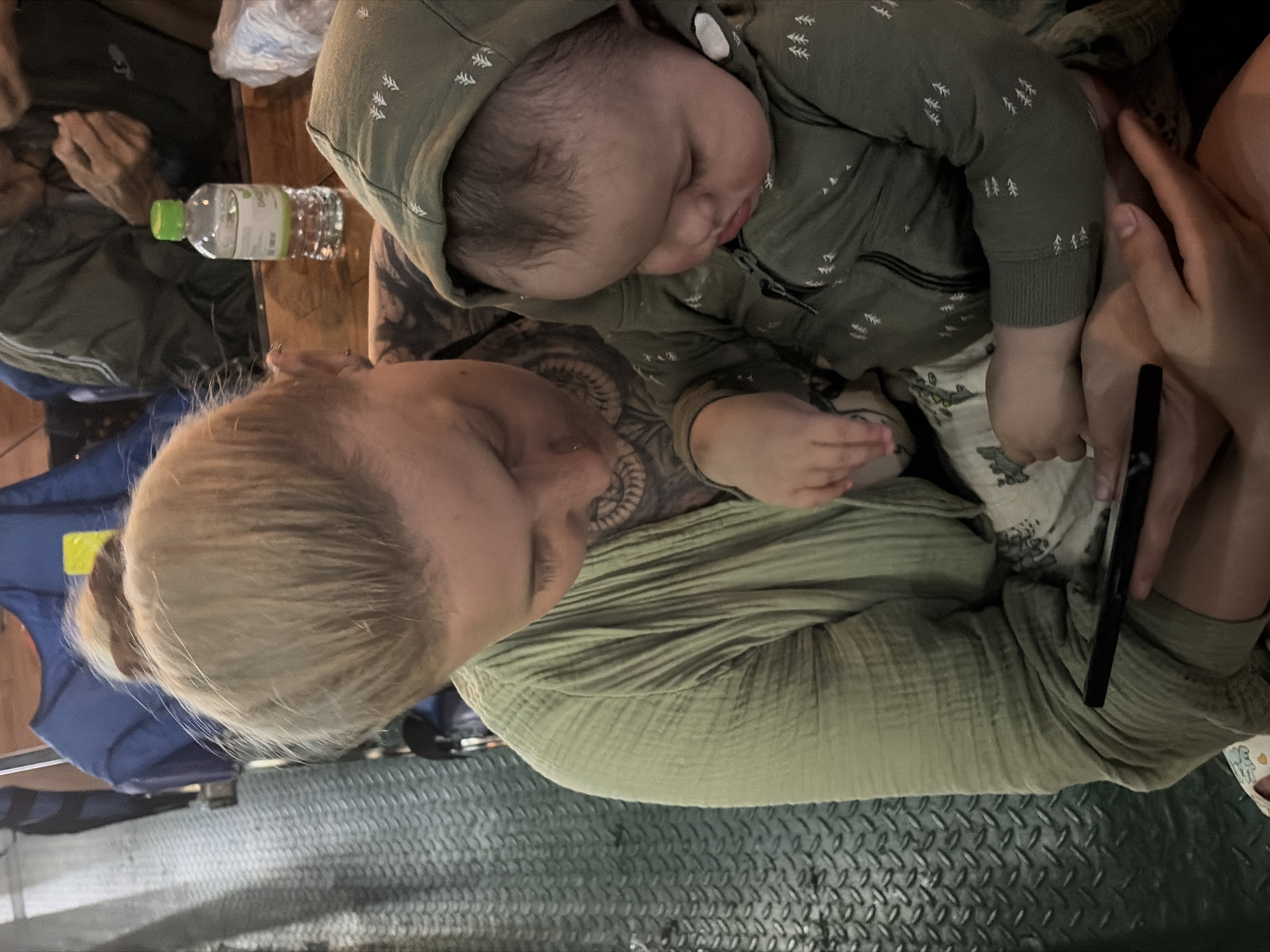 This image captures a tender moment between a woman and a baby in what appears to be an indoor or covered setting in Đà Nẵng, Vietnam.
The primary subjects are a woman, likely a mother, and an infant. The woman has blonde hair, pulled back, and visible tattoos on her arm and shoulder/chest area. She is wearing a light green, textured top, possibly made of muslin. The baby is nestled against her, appearing asleep or resting peacefully, dressed in a dark green hooded romper adorned with small white tree patterns. The woman's head is tilted downwards, and she is holding a dark-colored smartphone in her right hand, while her left hand is near the baby. Partially visible in the background, to the left, are the hands and arm of another person.
The setting features a dark, textured metal floor with a diamond plate pattern, which could suggest a ferry, boat, or a specific type of industrial-style establishment. A wooden surface, possibly a table or bench, is visible behind the woman and baby. On this wooden surface, a clear plastic water bottle with a bright green cap and a white label is placed.
The lighting is dim, indicating it might be evening or an interior with subdued illumination. The clothing choices—a lightweight top for the woman and a romper for the baby—suggest a warm climate, consistent with Đà Nẵng. The baby's serene expression conveys a sense of peace and comfort.
On the water bottle's label, faint text is visible, including what looks like "Nước Tinh Khiết" (Vietnamese for "Purified Water"), a common phrase on water bottles in Vietnam.
