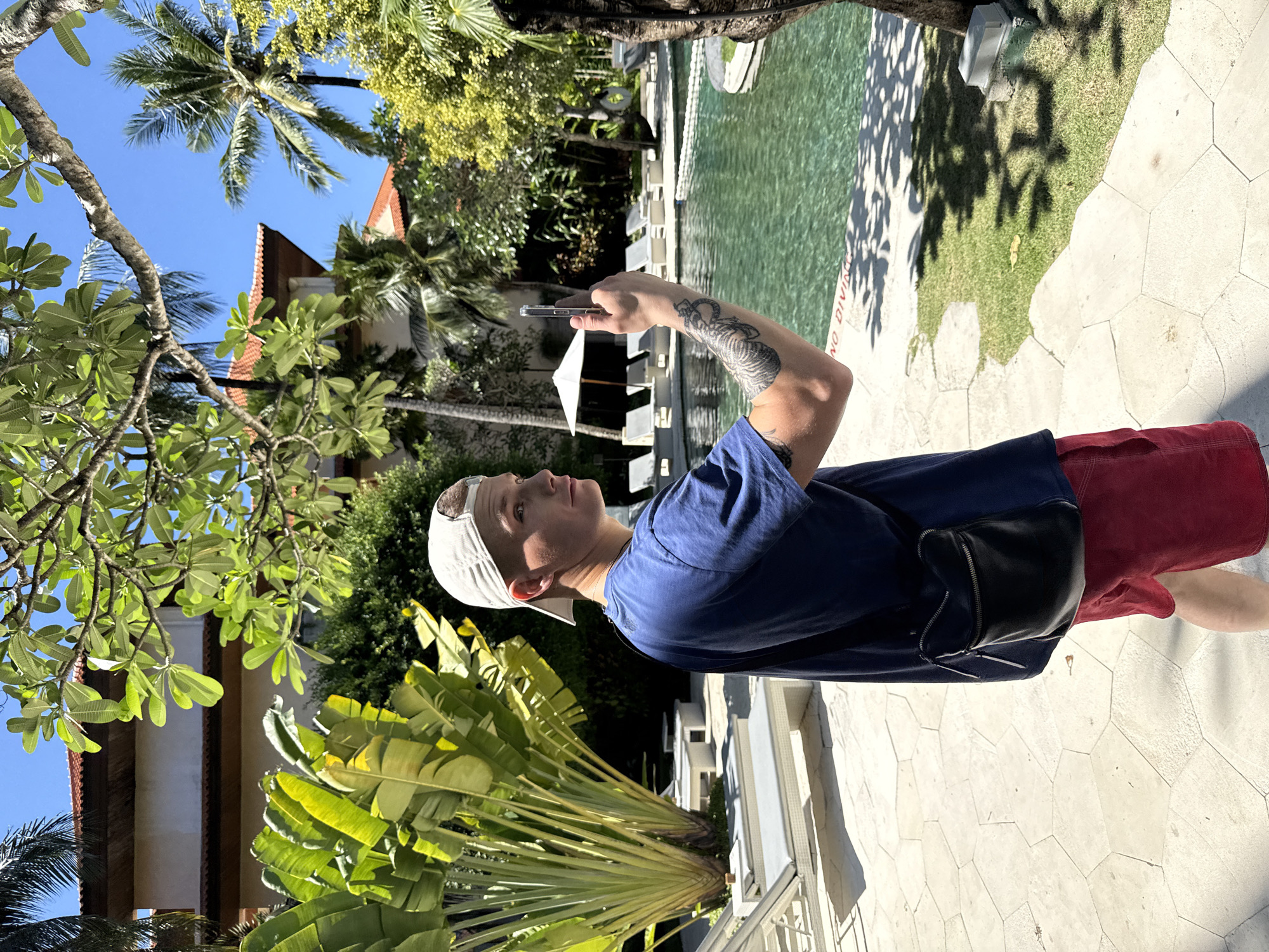 The image is a medium shot of a man standing near a swimming pool in an unknown location in Indonesia. The man is wearing a blue t-shirt, red shorts, and a white baseball cap. He has a black fanny pack slung over his shoulder and a tattoo on his left arm. He is holding a smartphone up in his left hand, as if taking a photo or video.
Behind the man is a swimming pool surrounded by white lounge chairs and umbrellas. The pool appears to be part of a resort or hotel complex. Lush green trees and palm trees are visible in the background, with glimpses of buildings peeking through the foliage. The sky is a clear, bright blue. The ground beneath the man's feet is paved with light-colored, irregularly shaped stones.