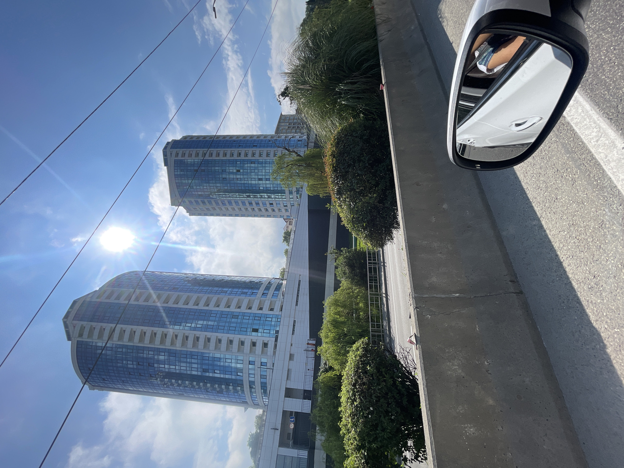 The image shows a view from a vehicle on a sunny day in Sochi, Russia. Two modern, glass-fronted residential buildings with white balconies are visible in the background. In the foreground, a concrete barrier lines the road, and the side mirror of the car reflects a person's arm and hand holding a phone. Lush green trees and bushes are planted between the road and the buildings. Overhead, power lines crisscross the bright blue sky, with the sun causing a lens flare. The image appears to have been taken from the coastal highway in Sochi, possibly near the port area or the Olympic Park, given the contemporary architecture.
