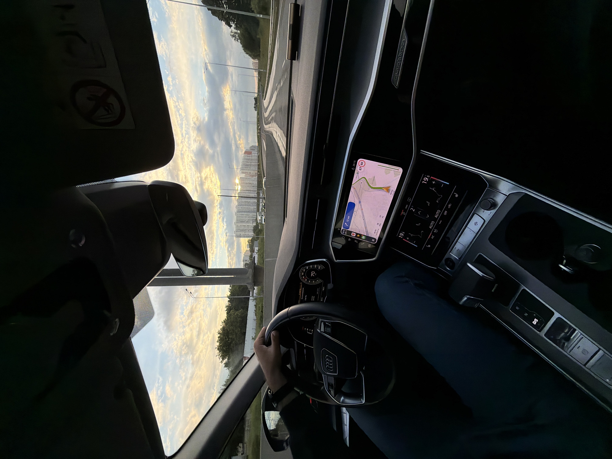 The image shows the interior of a car, viewed from the passenger seat, looking forward. The driver's hand is on the steering wheel. The dashboard features a digital instrument cluster displaying a speed of 70 and a large infotainment screen showing a navigation map. The car is on a highway, with a view of apartment buildings and trees lining the road. The sky is cloudy with hints of sunset. A sun visor is visible at the top of the windshield with a "no smoking" symbol. The transmission selector shows "D/S" and "R" indicators. The scene is consistent with a vehicle traveling on a highway in the city of Khimki, Russia, given the visual cues of the architecture and the highway infrastructure.