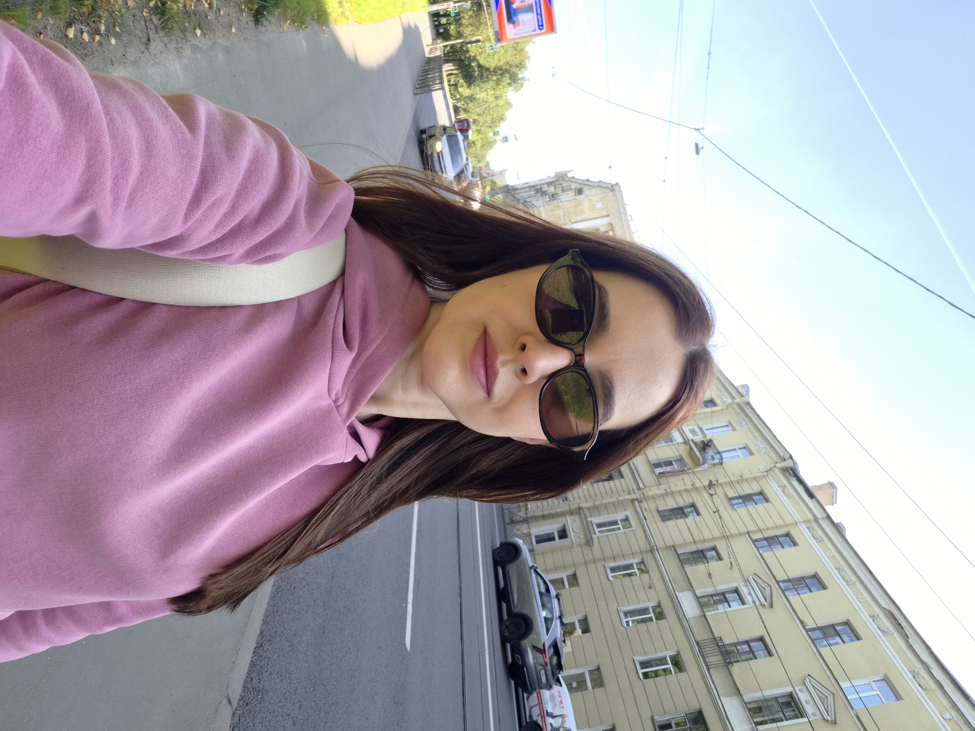 In a selfie taken on a sidewalk in Saint Petersburg, Russia, a woman with long brown hair and sunglasses wears a pink hoodie and a light-colored strap across her shoulder. Behind her, a building with yellow facades and numerous windows lines a city street. Cars are visible driving on the street, and overhead wires are strung across the sky. To the left, trees and a billboard are present. The sky is blue with some contrails visible.