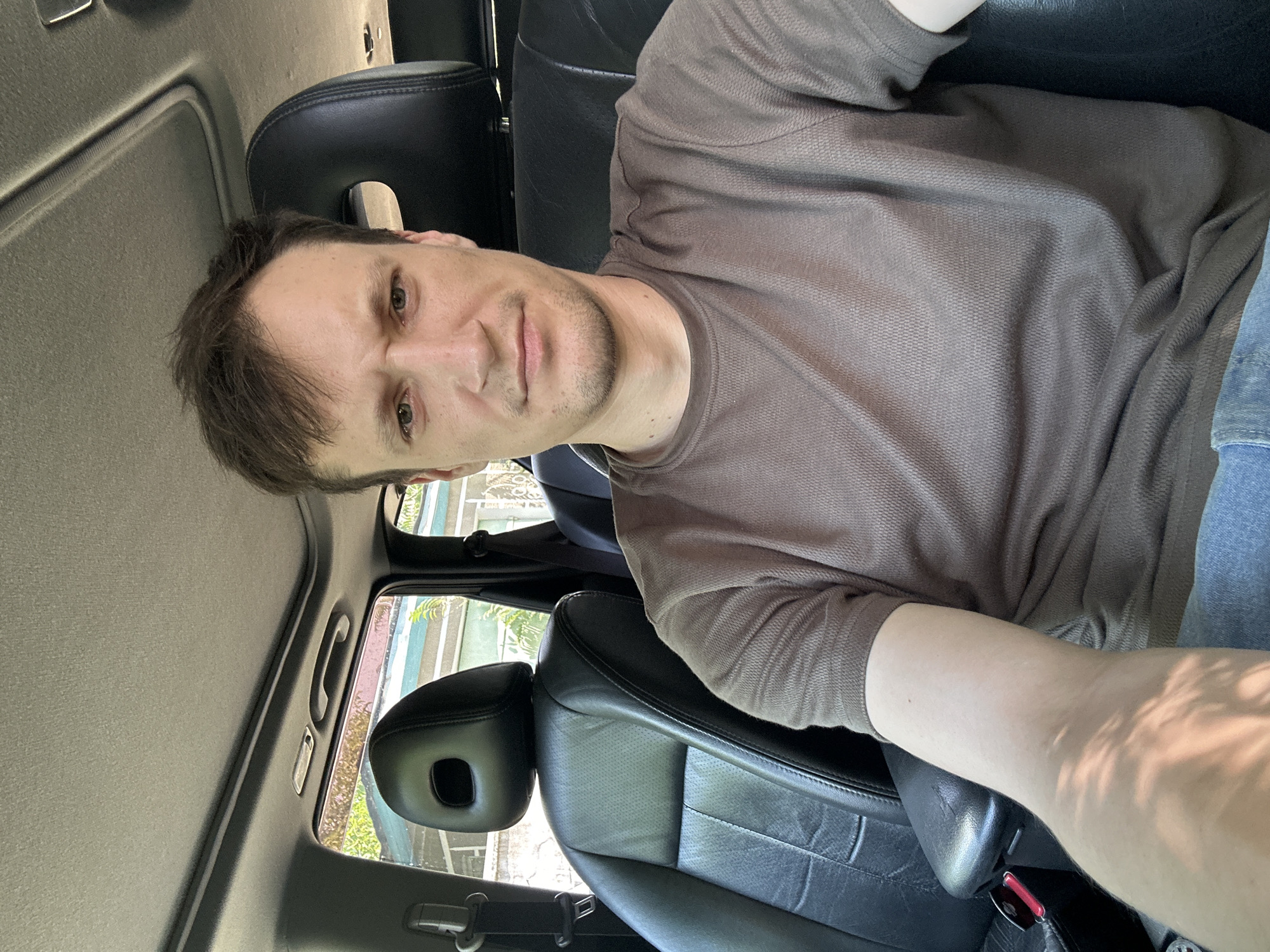 A man is seated in the passenger seat of a car. He is wearing a brown t-shirt and blue jeans. The car has black leather seats and a sunroof. The man is looking at the camera with a neutral expression. Outside the car, there are trees and buildings visible, suggesting an urban or suburban environment. The specific location is Makiv, Ukraine.
