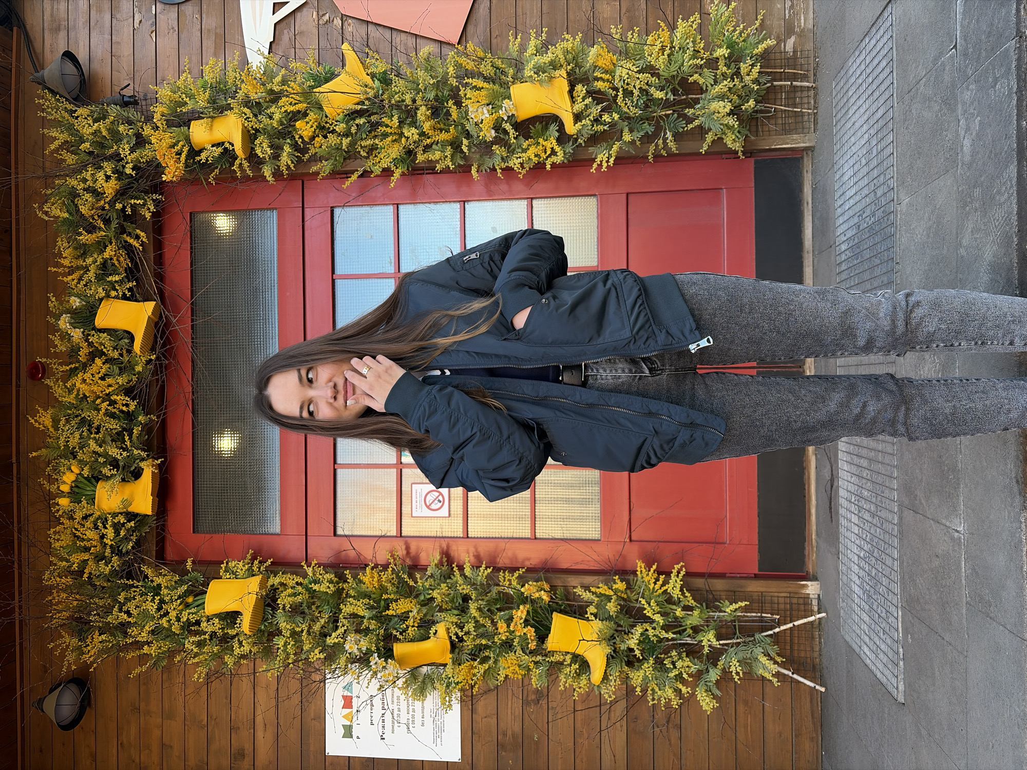 The image features a young woman standing in front of a red door decorated with yellow mimosa branches and yellow rubber boots, possibly as part of a decorative installation. The woman has long brown hair and is wearing a dark blue jacket and gray jeans. She has her hand near her face and is smiling. The setting appears to be outdoors, in front of a wooden-clad building. A sign with text in Russian, including the word "ресторан" (restaurant), suggests that the location is a restaurant. The text on the sign includes opening hours and indicates that the restaurant is open every day. The location is Izhevsk, Russia. The lighting is soft and diffused, indicating an overcast day. A gray concrete wall can be seen to the right of the door.