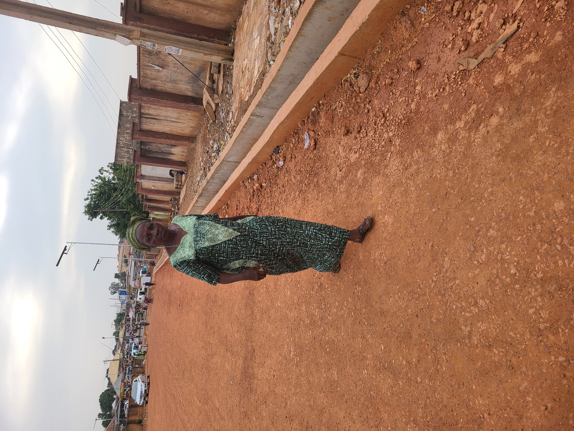 The image displays a full-body portrait of a woman standing on a reddish-brown dirt road, with a bustling street scene in the background. The woman, an adult, is positioned towards the right of the frame, looking directly at the camera with a neutral expression. She is dressed in a long, patterned green and black gown with long sleeves, and a matching green headscarf. She wears dark sandals on her feet.
To the woman's left, a concrete drainage channel runs parallel to the road. Beyond this channel are several simple, low-rise buildings with worn, reddish-brown walls and some exposed concrete elements. Utility poles with overhead wires are visible, and one pole has several pieces of paper attached to it, likely notices or advertisements, though the text is illegible.
The road itself is unpaved, made of reddish-brown soil and gravel. Further into the distance, the road is lined with more buildings and trees, and a variety of vehicles including cars and what appear to be auto-rickshaws or tricycles, indicating a busy street or market area. The sky is overcast, suggesting daytime. The landscape, with its distinctive reddish-brown earth and urban characteristics, aligns with the context of Jos, Nigeria.