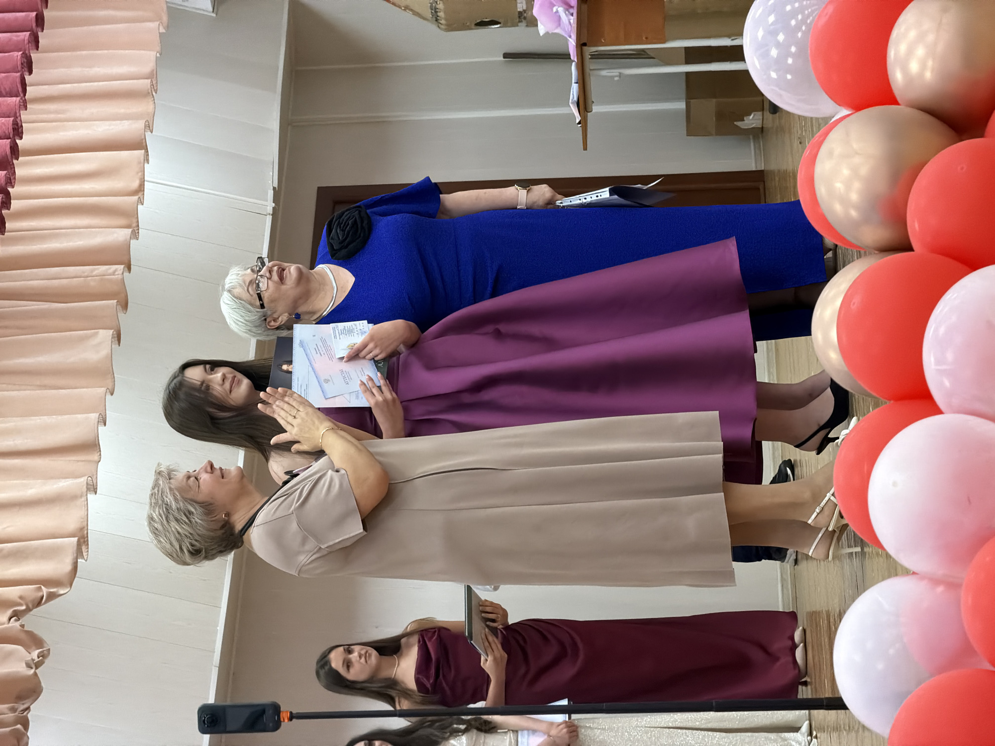 The image depicts a celebratory gathering, likely a graduation or awards ceremony, taking place indoors. Several individuals, predominantly women, are present, dressed in formal attire. The central figures appear to be receiving or presenting certificates or awards, indicated by the documents held in their hands. One woman in a vibrant blue dress is smiling and holding a certificate. Beside her, a woman in a beige dress looks up with an engaged expression. Further down, another woman in a burgundy dress is also holding a document.
The setting features decorative elements commonly associated with celebrations, most notably a cluster of balloons in red, white, and gold. The background includes what appears to be classroom or auditorium furniture, including a wooden shelf and curtains. The lighting suggests an indoor environment, possibly during the daytime given the overall brightness.
The mood appears joyful and accomplished, with expressions of happiness and engagement on the faces of the people. The presence of formal wear and awards strongly suggests a formal occasion. The scene is vertical, with the subjects oriented as if standing. No specific time of day or weather conditions are discernible. While the documents held by the individuals show some text, it is not clearly legible to be transcribed. The location context is Moscow, Russia, which does not introduce any visually specific cues in this particular image.