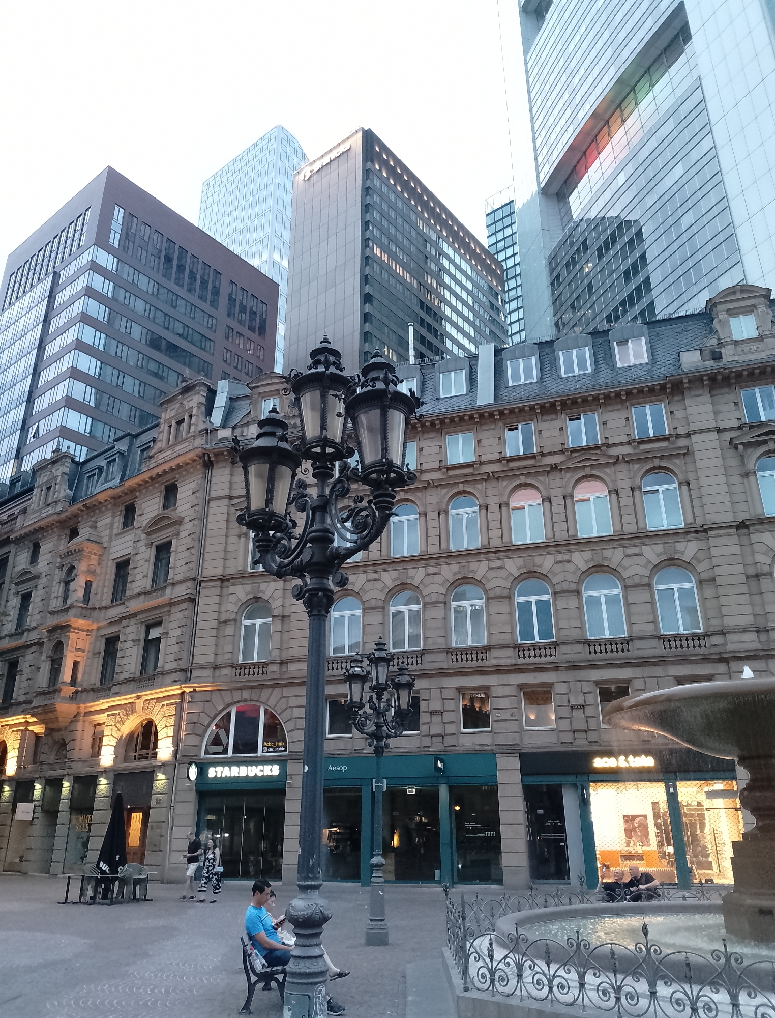 In a city square, an ornate lamppost stands in the foreground, while in the background, modern skyscrapers loom over a historic building. A Starbucks storefront is visible on the ground floor of the historic building, and a man sits on a bench, looking at his phone. To the right, a fountain adds a touch of tranquility to the urban landscape. The sky is overcast, suggesting a possibly cool or temperate climate.
