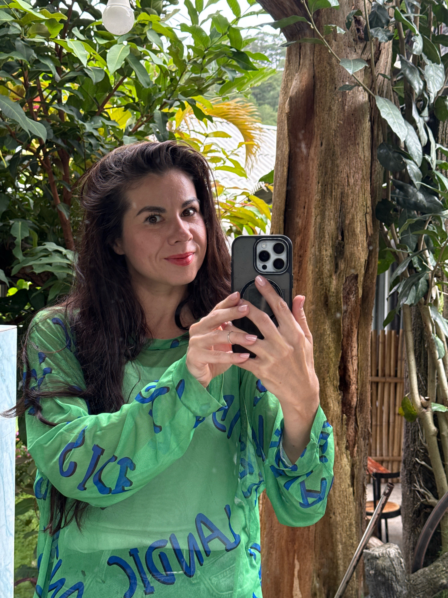 A woman with long, dark hair and light skin takes a selfie with her smartphone. She is wearing a sheer green long-sleeved shirt with large blue letters that appear to spell out "ICE" and "SIGNAL." Her left hand is visible, wearing a silver ring. The background suggests an outdoor environment with lush green foliage and a large tree trunk. The presence of a lightbulb hanging from a wire and what appears to be a covered structure in the background might indicate a greenhouse or a covered outdoor seating area. The woman is smiling slightly as she captures the photo. The scene is likely located in Hiệp Thạnh, Vietnam, given the surrounding vegetation and potentially the architectural elements visible.