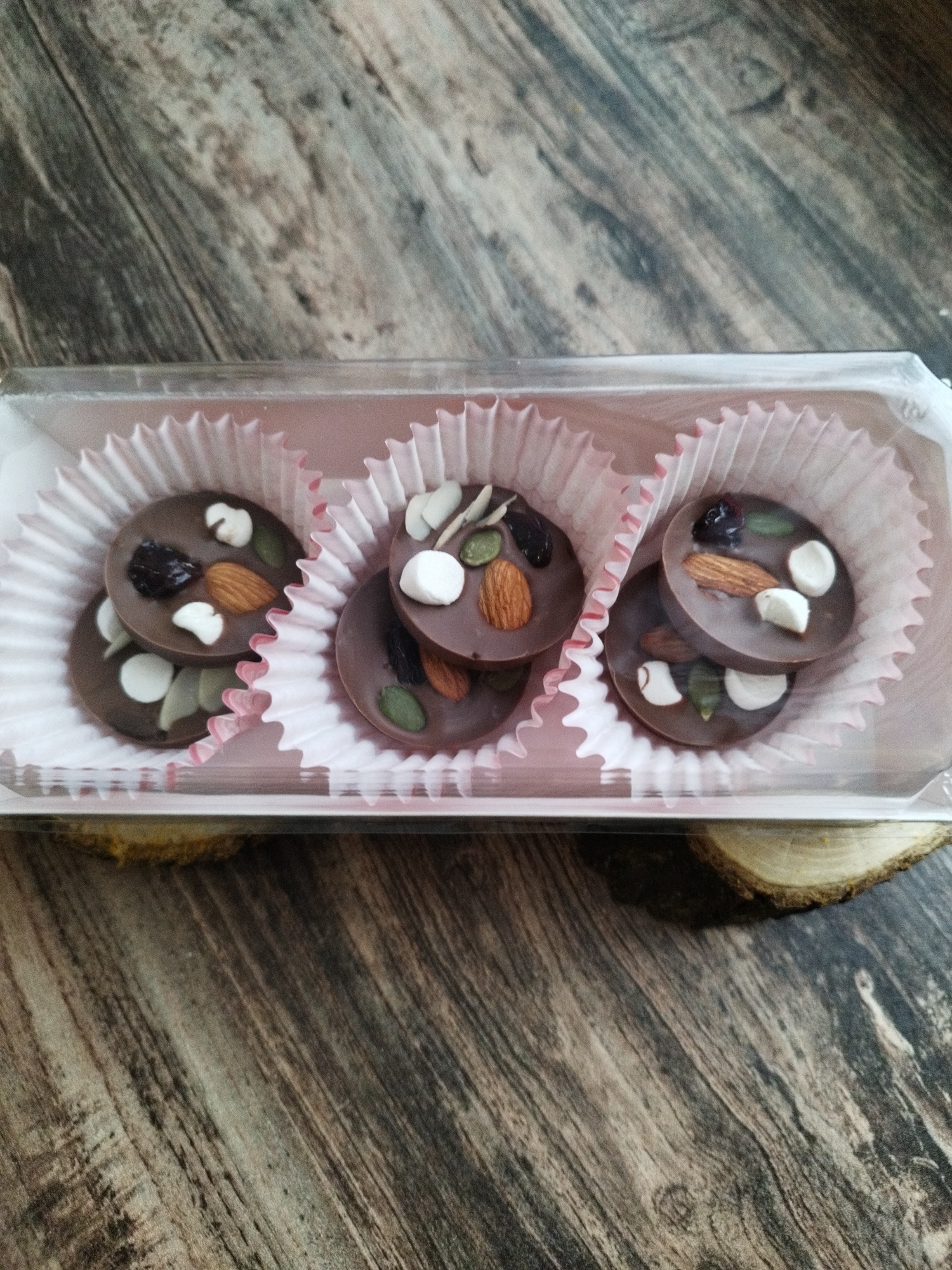 A clear plastic container holds three pink paper cupcake liners, each containing two stacked chocolate discs. The chocolate discs are dark brown and adorned with various toppings including what appear to be slivered almonds, dried cranberries or raisins, pumpkin seeds, and white pieces that could be marshmallows or white chocolate chips. The background is a wooden surface with a distressed, grayish-brown grain pattern. The image is taken from a slightly elevated angle looking down at the chocolates. The location is not specified in the image, and thus, cannot be provided.