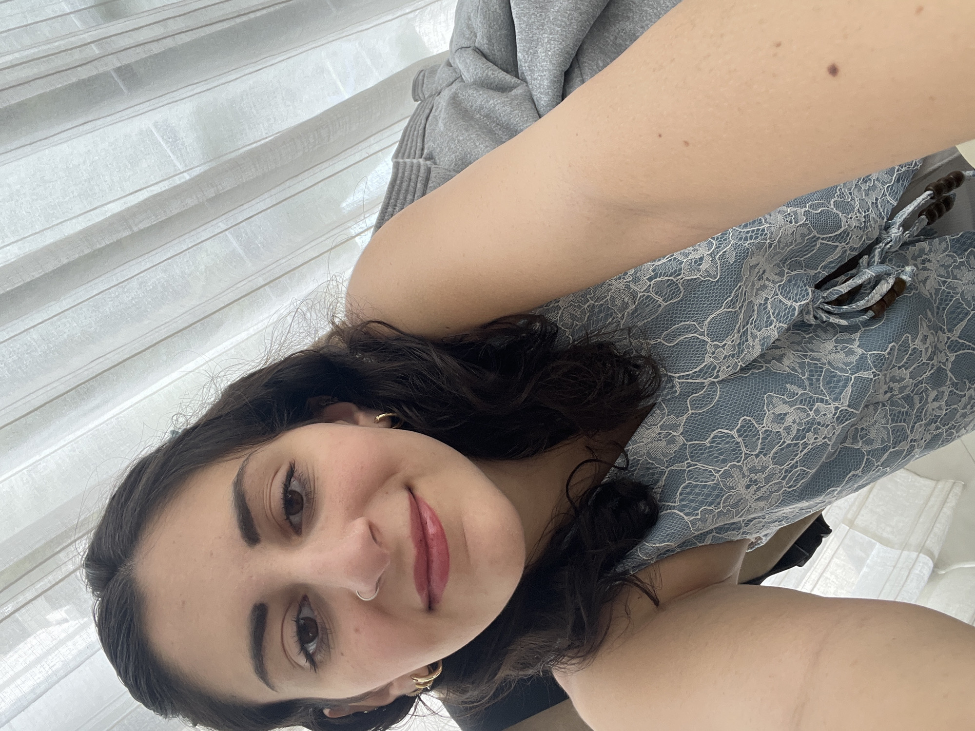 A selfie-style photograph depicts a woman indoors. She has dark, wavy hair, brown eyes, and fair skin. She is wearing gold hoop earrings and a nose ring. Her lips are painted with a red lipstick.
Behind the woman is a grey hoodie, and a light blue garment with a white lace pattern. In the background, white curtains can be seen.
There is no information in the image to ascertain that it was taken in Mexico City, Mexico.