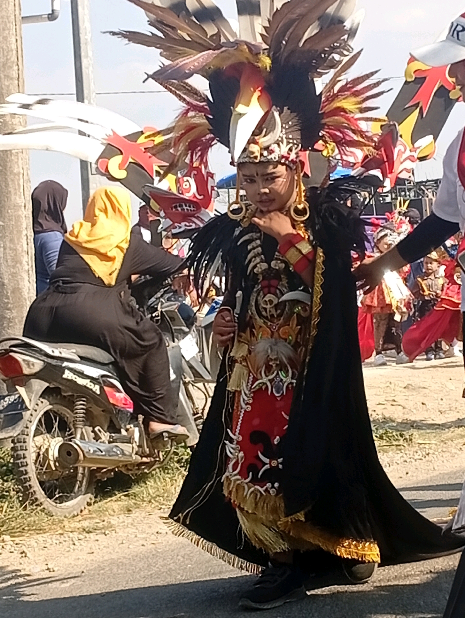 This image depicts a vibrant street scene in Nglojo, Indonesia, likely during a festive event or parade. In the foreground, a young person adorned in an elaborate, traditional costume with a large headdress featuring feathers and avian-like elements stands prominently. The costume is detailed with gold accents, black fabric, and red embellishments, suggesting a cultural performance. To the left of the foreground figure, a woman wearing a bright yellow hijab sits on a motorcycle, dressed in a long black garment. Behind her, another woman in a dark hijab is visible. In the background, other individuals are present, some also in colorful attire, and there are indications of stalls or structures that might be part of the event. The ground is a mix of asphalt and gravel, with dry vegetation visible on the sides. The lighting suggests it is daytime, under a clear sky.