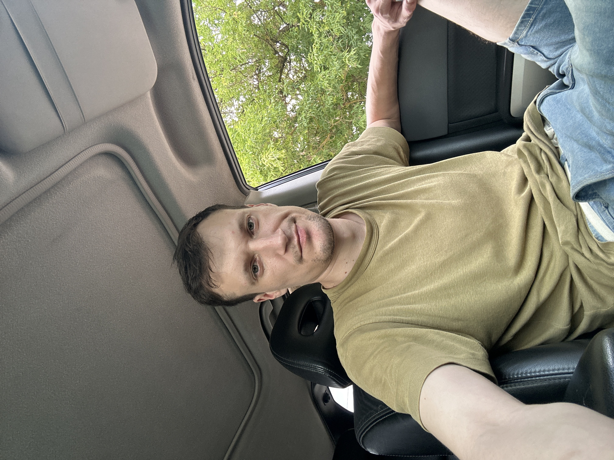 A man is sitting in the driver's seat of a car, looking at the camera. He has short brown hair and is wearing a tan t-shirt and denim shorts. The car's interior is visible, including the black leather seats and the sunroof. Outside the car, green trees are visible through the window. The image appears to have been taken during the day. The specific location within Makiv, Ukraine is not discernible from the image itself.