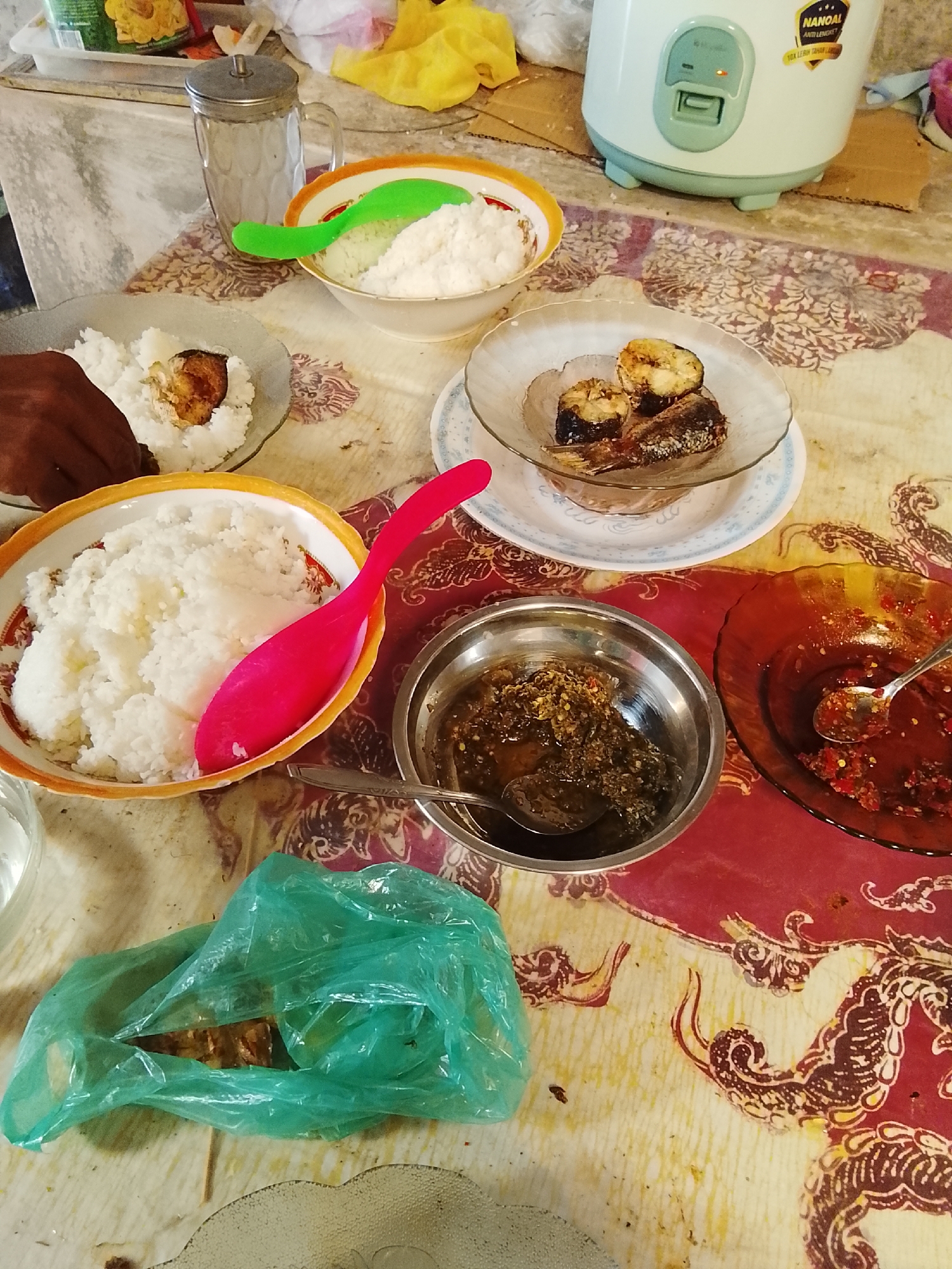 This is a top-down, medium shot of a casual meal set on a table with a patterned tablecloth. In the foreground, a green plastic bag contains what appears to be food. To the left of the bag, a clear glass contains a beverage. A large, orange-rimmed bowl filled with white rice is prominent, with a bright pink serving spoon resting inside. To the left of this bowl, a hand is reaching into another bowl containing rice and a piece of food, possibly fried. Behind the rice bowls, there is a glass jug with a metallic lid. A smaller, clear bowl with three pieces of fried fish sits on a white plate with a blue rim. Further to the right, there are two small, dark brown bowls containing sauces or relishes, one with a spoon. A glass rice cooker is visible in the background, with a label that reads "NANOAL ANTI LENGKET 10x LEBIH TAHAN LAMA". The scene suggests a home-cooked meal in a rustic or informal setting. The location is identified as Campago Barat, Indonesia.