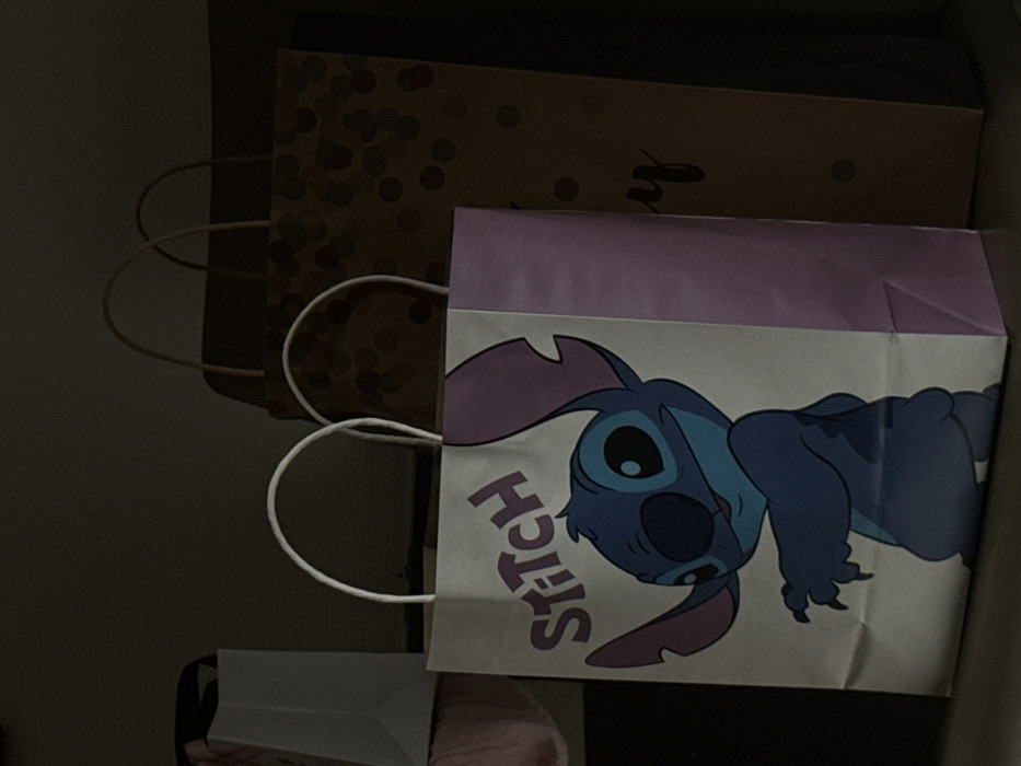 This scene, situated in the city of Albi, France, depicts an indoor, dimly lit environment featuring multiple paper gift bags arranged on a flat surface. In the immediate foreground, a white and light purple gift bag stands upright. This bag features a prominent graphic of the blue Disney character Stitch, with large ears, and the word "Stitch" printed in purple above its head. To the right of and slightly behind this primary bag, a taller, darker brown gift bag is visible. It is patterned with numerous dark circular dots and exhibits partial black cursive text, specifically the letter "y," on its lower right side. To the far left, a portion of another white gift bag and an indistinct reddish-pink or light brown object are partially visible in shadow. No persons are present within the frame, and no actions or interactions are depicted beyond the static arrangement of the objects.