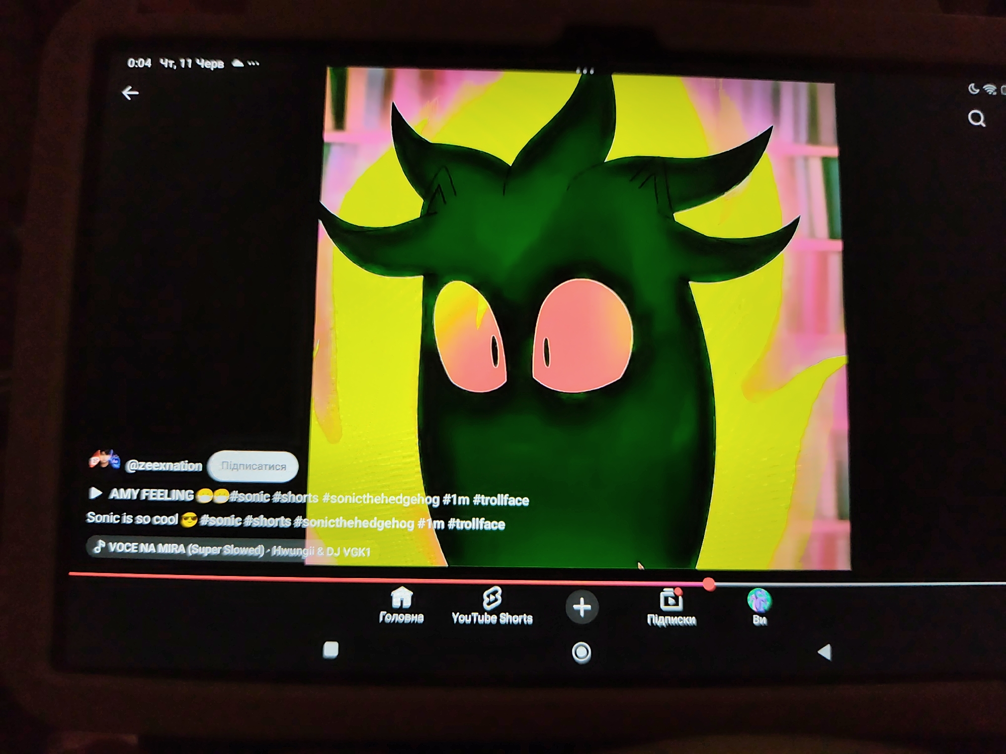 The image shows a screenshot of a YouTube Shorts video playing on a tablet. The video features a cartoon character with spiky green hair and large pink eyes against a bright yellow and pink background. The video is hosted by a user named "@zeexnation" and includes hashtags like #sonic, #shorts, #sonicthehedgehog, #1m, and #trollface. Below the video title, there is a music track listed as "VOCE NA MIRA (Super Slowed) - Hwungii & DJ VGK1". The tablet's interface shows navigation options for "Home," "YouTube Shorts," "Subscriptions," and "You." The time displayed at the top of the screen is "0:04 Чт, 11 Черв" which translates to "0:04 Thursday, 11 June." The tablet is likely located in an indoor setting, and no specific landmarks of Kryvyi Rih, Ukraine, are visible.