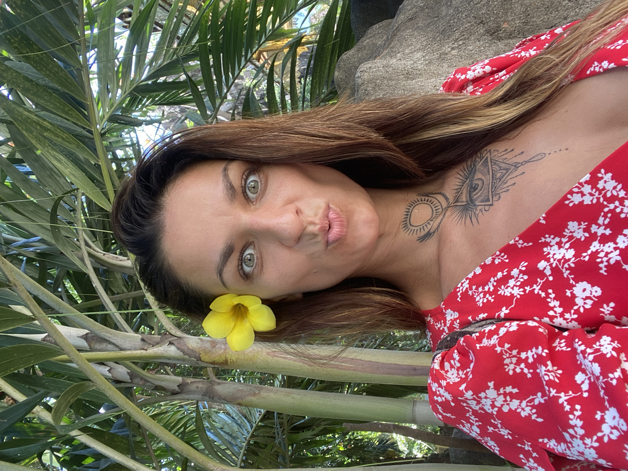 A woman is pictured outdoors, among tropical foliage. A yellow flower is placed in her hair near her ear. She is wearing a red dress with a white floral pattern. Tattoos are visible on her neck and chest. The location is unspecified, and there is no indication it is in Dĩ An, Vietnam.