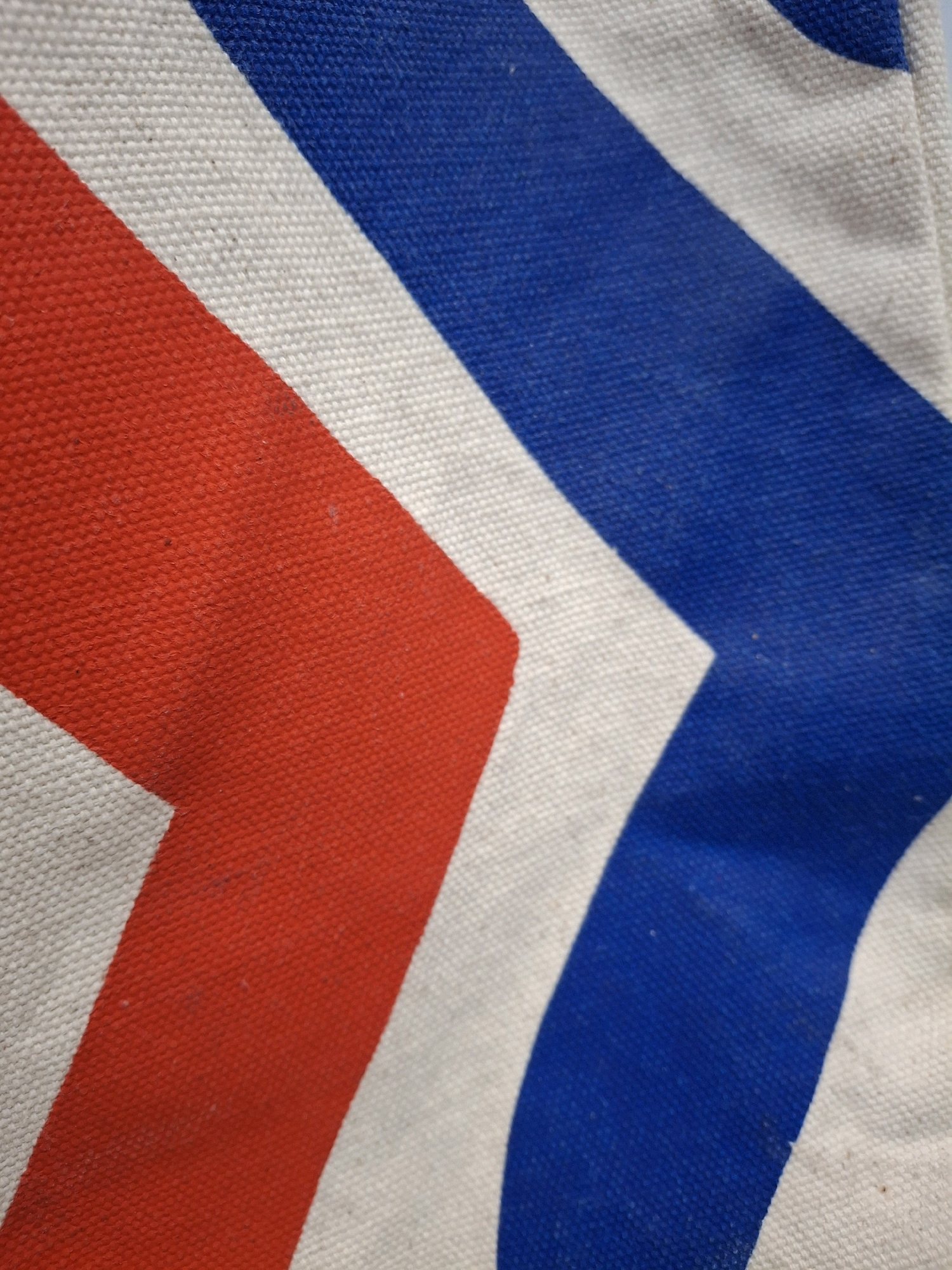 This is a close-up, oblique shot of a canvas material with diagonal stripes of red, white, and blue. The texture of the canvas is visible, and there are some faint smudges on the white and blue areas. The red stripe on the left appears to be a solid, bold color, while the blue stripe on the right is also a vibrant shade. The white stripes serve as a background between the colored bands. The image does not contain any elements that can be used to determine its location in the city of Lumajang, Indonesia, or any specific environmental or human context.