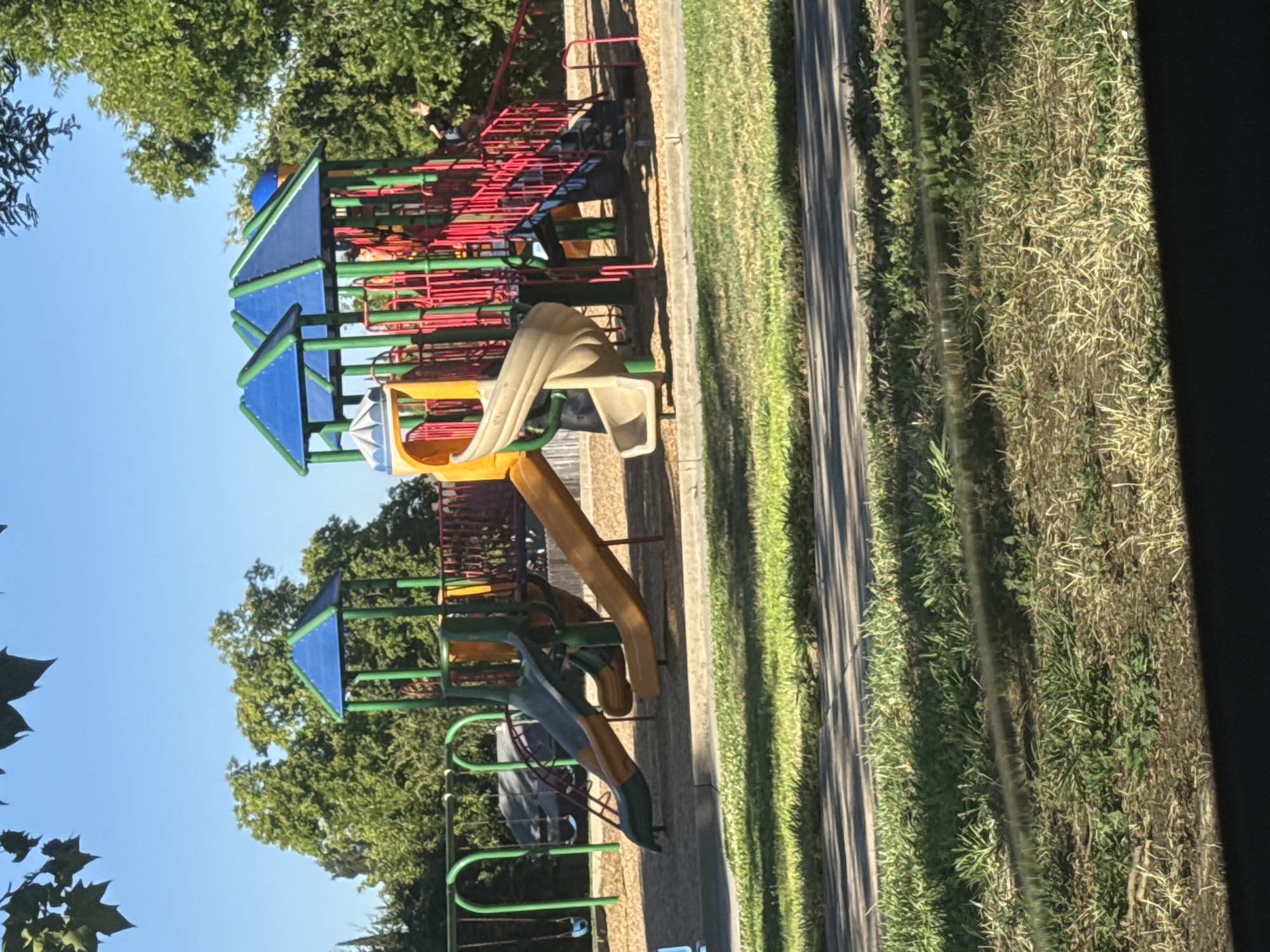 The image depicts a colorful playground structure with multiple slides and swings, situated outdoors. The playground features a main structure with red railings, blue and green roofs, and yellow and green slides. Adjacent to it, there is a swing set with green frames. The ground beneath the playground is covered with wood chips. The scene is set in a park-like environment with lush green trees and a clear blue sky overhead. There is a paved pathway and a grassy area in the foreground, with shadows cast by trees. The image is taken from a slightly elevated perspective, looking towards the playground. The location is identified as a park in Sacramento, United States. There is one visible person, a child, partially visible on the right side of the playground structure, seemingly interacting with it.