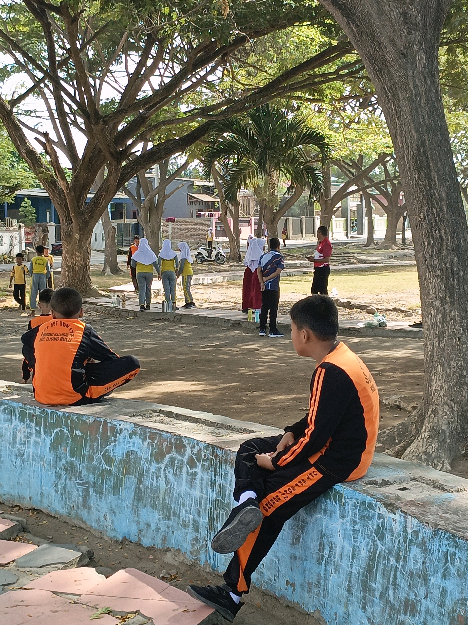 The image depicts an outdoor scene with several individuals, primarily children, in what appears to be a school or recreational area. In the foreground, two boys are seated on a low, weathered blue wall. The boy on the left, viewed from the back, wears an orange and black long-sleeved shirt with writing on the back and black pants. The boy on the right, also facing away, is dressed in a similar orange and black outfit, with orange stripes on the sleeves and pants.
In the middle ground, several children are walking and standing. Some are wearing yellow shirts and grey pants, with some girls also wearing white headscarves. A man in a red shirt and black pants is standing and talking to a person wearing a blue and black shirt and red skirt. Further in the background, there are buildings and more trees. A white motorcycle is parked on the right side of the background. The environment is characterized by large trees with spreading branches and a dusty, unpaved ground. The overall atmosphere suggests a warm, sunny day. This scene is likely located at a public space or educational institution within the city of Bulukumba, Indonesia, possibly near the coast given the presence of palm trees and the general tropical climate implied by the attire and vegetation. The writing on the boy's shirt, "UPT. SPF SD N 172," suggests it's associated with an elementary school.