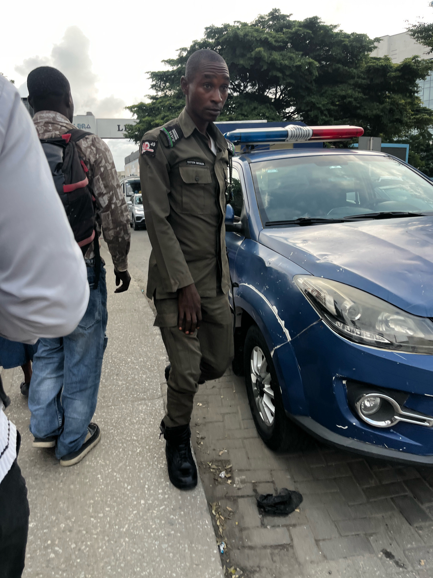 A uniformed Nigerian police officer stands on a paved sidewalk next to a blue police vehicle in Lekki Phase I, Nigeria. The officer is wearing a dark green uniform with patches on the sleeves and a name tag that reads "TITUN MORIW". He has dark skin and short black hair. His left hand is partially visible near his pocket, and his right hand is bent at the elbow. The police vehicle, a blue sedan, has a light bar with red and blue lights on its roof and appears to have some damage to its bodywork, including scuff marks and dents. A second individual, wearing a camouflage-patterned shirt and a backpack, stands behind the police officer, with his back to the camera. Another person, whose head and shoulders are visible on the left of the frame, is wearing a white top. The environment suggests an urban setting with trees and buildings in the background. The sidewalk is made of interlocking bricks, and there is some debris on the ground. The sky appears overcast.