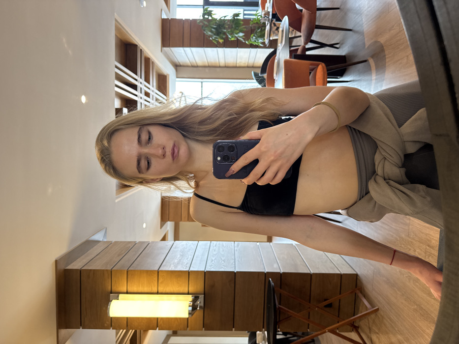 A woman with long blonde hair holds a dark smartphone, presumably taking a mirror selfie. She wears a thin-strapped black top. A light brown sweater is tied loosely around her waist. She seems to be in a modernly decorated space, potentially a hotel or apartment. The background includes wooden paneling, a lighted wall fixture, and a glimpse of a dining area with chairs and a plant. The perspective is slightly skewed.