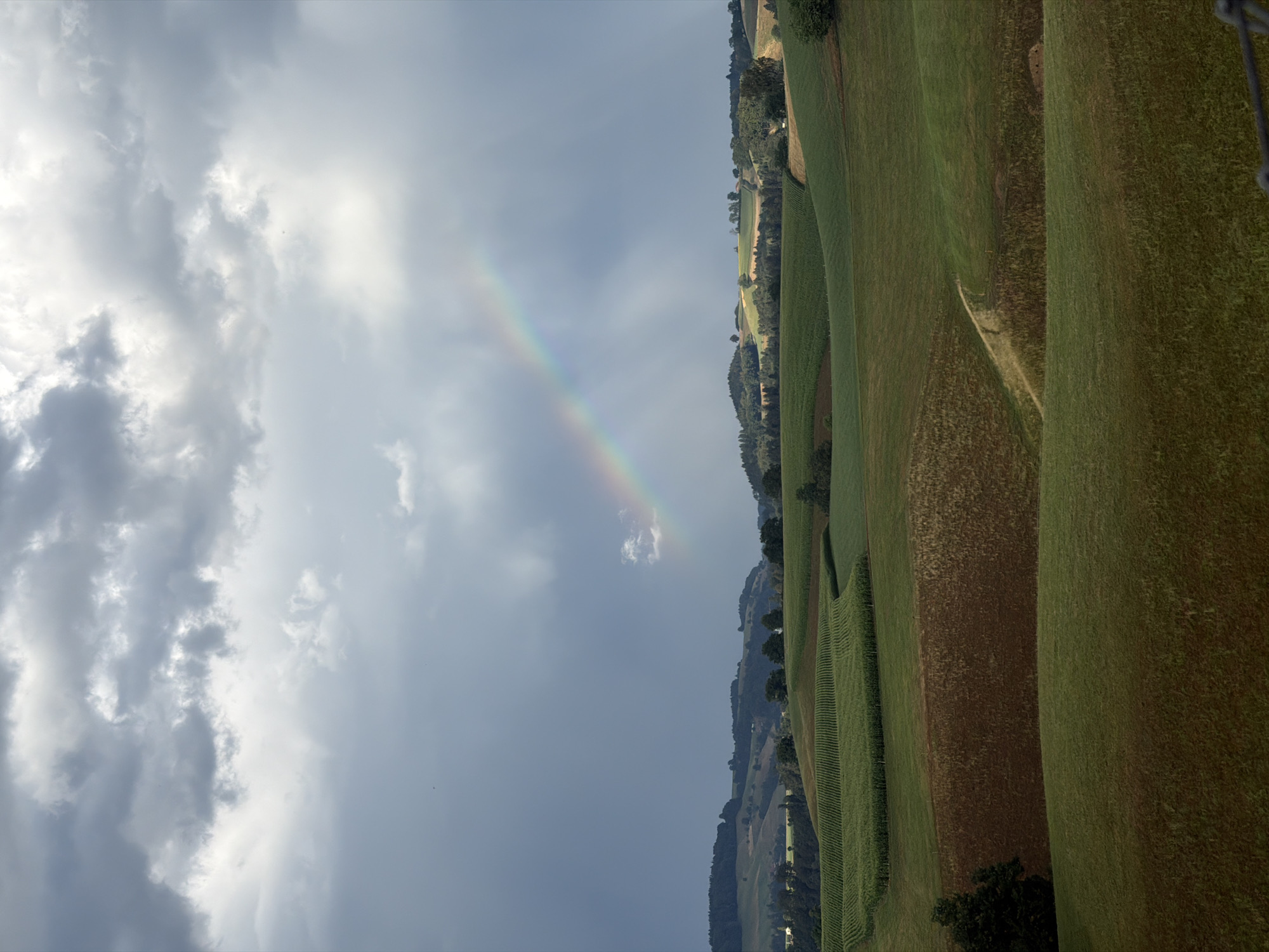 This is a vertical, eye-level, full shot photograph of a rural landscape in Ufhusen, Switzerland. The image features rolling green hills covered in fields of crops and pasture. A faint rainbow arcs across the sky, appearing as a colorful band against the dark, stormy clouds. The foreground is dominated by lush green fields, with a path winding through the middle ground. In the background, a treeline separates the farmland from the sky. The overall mood is serene and tranquil, with a hint of drama from the stormy sky. There are no people or animals visible in the image.