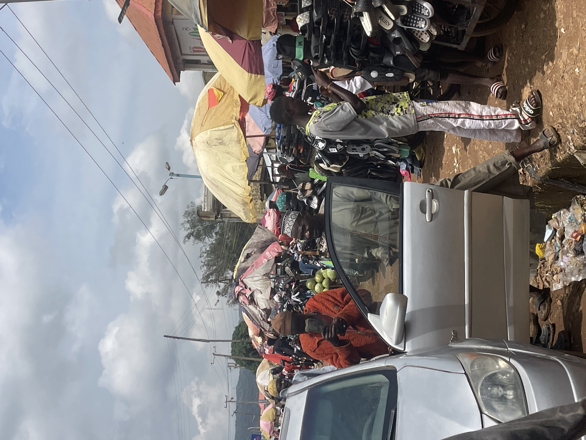 The image depicts a bustling street market scene in Rafin Par, Nigeria. The foreground features the driver's side of a silver sedan with its door open, revealing the lower body of a person seated inside. To the left, partially visible, is a person wearing a dark garment.
The central focus is on several men interacting within the market. A man in an orange jacket and brown cap is holding what appears to be a phone or wallet up to his face, seemingly examining it. He is positioned between the car and a stall. To his right, another man in a light-colored shirt and patterned trousers is looking towards a display of footwear. He is wearing black and white striped sandals. Further to the right, a man in a light green garment and a white cap is visible.
The market stalls are covered by a variety of umbrellas, some weathered and torn, displaying a diverse assortment of goods, most notably a large quantity of footwear arranged on shelves and racks. In the midground, there are several large green fruits, likely gourds or melons, piled on the ground.
The environment is outdoors, under a cloudy sky. Overhead power lines are visible, as are streetlights and distant trees. The ground appears to be a mixture of dirt and debris, with trash scattered in some areas. The overall atmosphere suggests a busy, commercial hub.