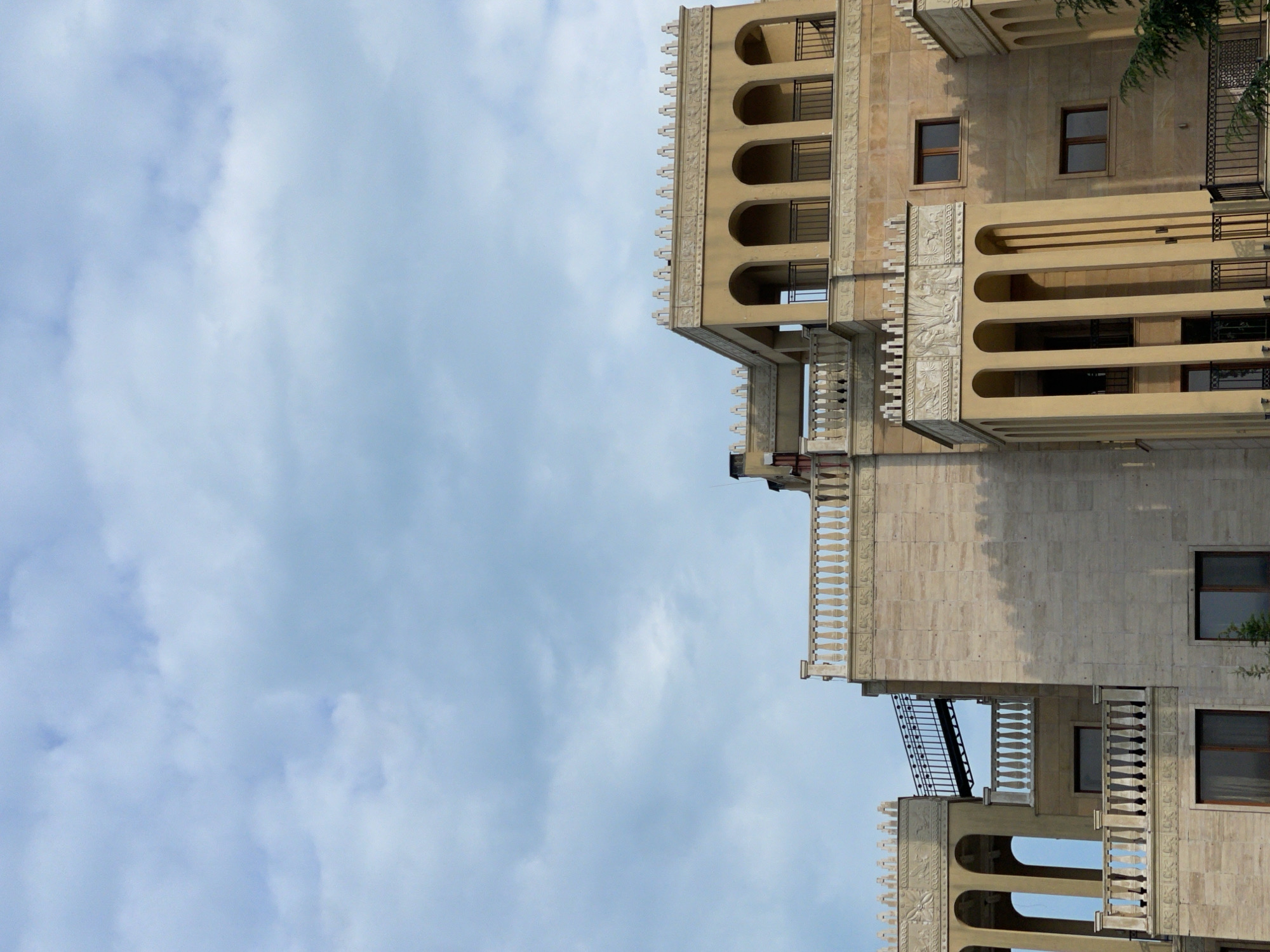 The image, originally rotated 90 degrees counter-clockwise, depicts a section of an ornate, multi-story building set against a cloudy sky. When viewed upright, the building occupies the right side of the frame, showcasing its architectural details. It is constructed from a light-colored stone or stucco, featuring a classical or Art Deco-inspired style.
Prominent architectural elements include several balconies: some are deep, recessed arches with dark metal railings, while others feature intricate white balustrades. Decorative carved friezes adorn the facade, particularly along cornices and between levels, adding to the building's grand appearance. Multiple rectangular windows with dark frames are visible across the structure. A small amount of dark green foliage is present on the far right, partially obscuring the building.
The left and upper portions of the frame are dominated by a pale blue sky filled with soft, diffused white and light grey clouds, indicating an overcast daytime. There are no people or explicit activities taking place, and no visible text. The scene is a static architectural study located in Varna, Bulgaria.