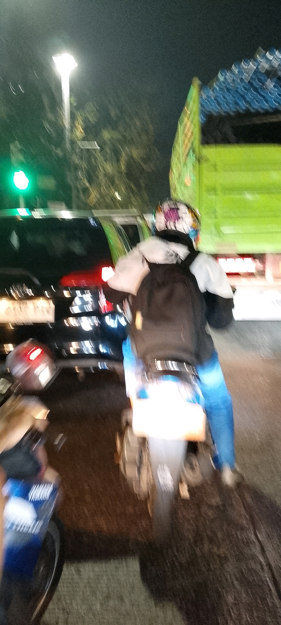 In the city of Bekasi, Indonesia, at night, a person is riding a motorcycle. The person is wearing a helmet and a backpack. The motorcycle is in motion, and the background is blurred, indicating movement. Several vehicles are visible, including a truck and a car. A traffic light, appearing green, is also present in the scene. The overall impression is a busy street at night.
