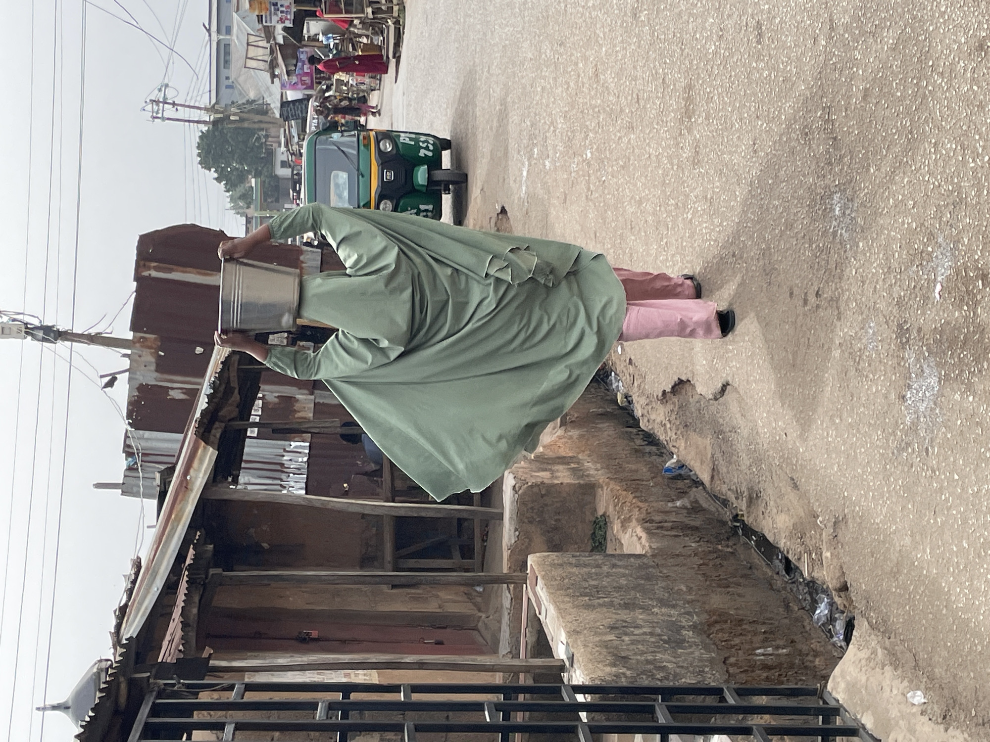 A person wearing a green robe and pink pants is carrying a metal bucket on their head while walking on a paved street in Jos, Nigeria. The person is walking past a building with a rusted corrugated metal roof and a black metal fence in the foreground. Behind the person is a green and yellow auto rickshaw. In the background, there are other people and buildings. The sky is overcast.