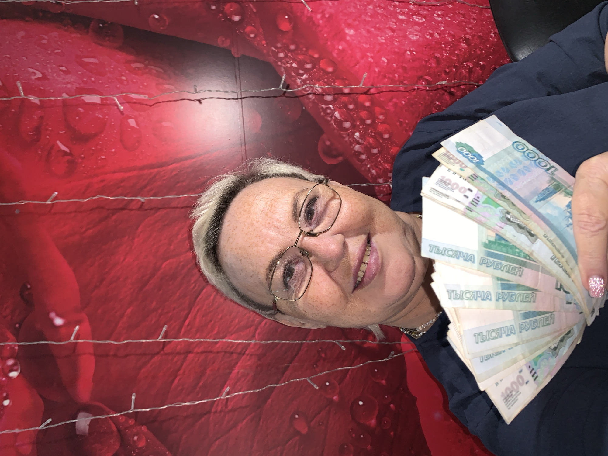 A medium shot shows a Caucasian woman in her late 50s or early 60s, with short blonde hair and freckles, holding a fan of Russian rubles in her right hand. She is wearing glasses and a dark blue top. Her left hand is not visible. The background is a red textured surface with water droplets and a string of small lights. The image appears to be a selfie. The location is not discernible in the image.