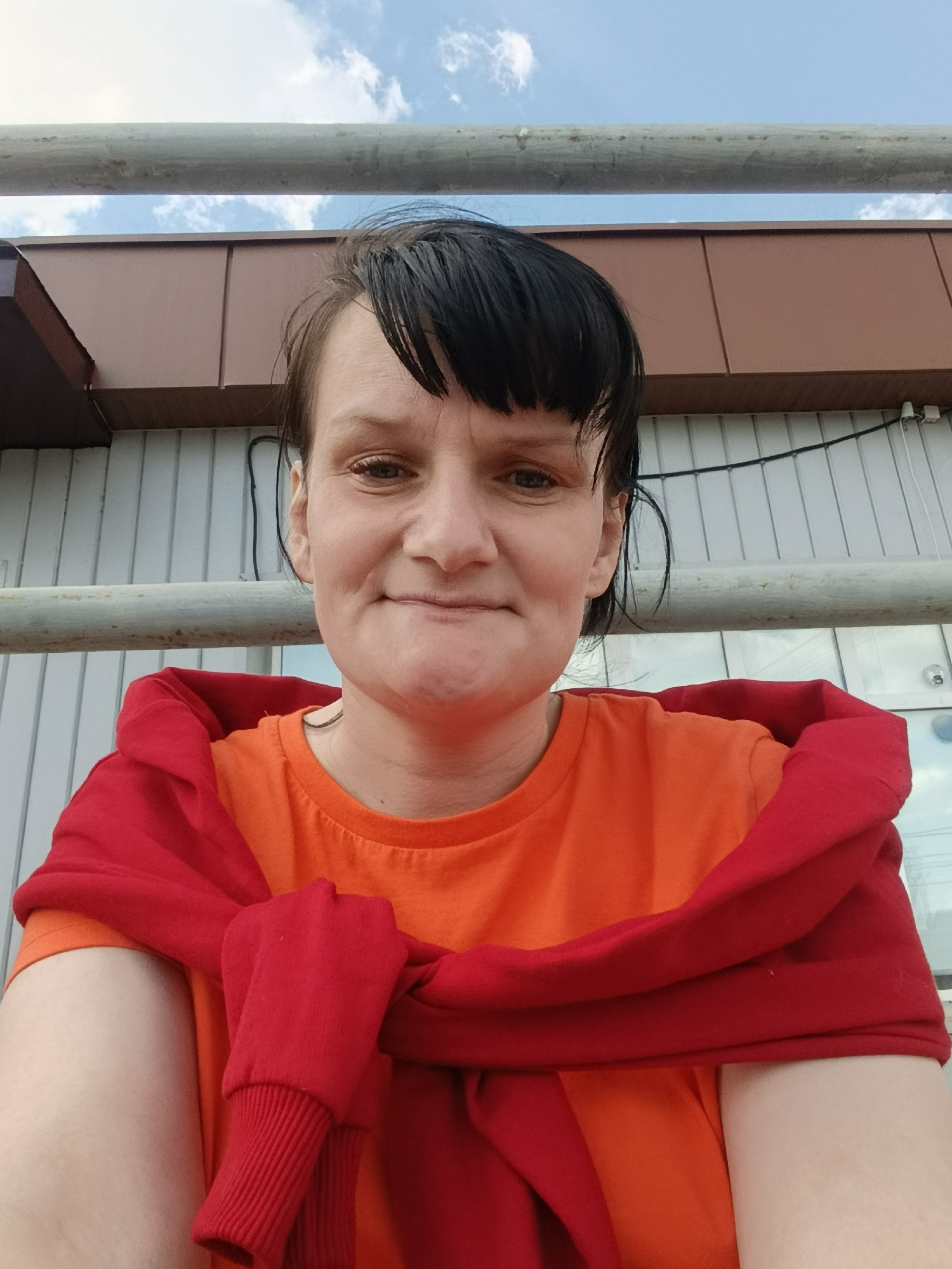 A woman is posing for a selfie outdoors. She has dark hair with bangs, fair skin, and is wearing an orange t-shirt with a red sweater tied around her shoulders. Behind her is a building with brown trim and a white, vertically-lined facade, along with a grey metal railing. The sky is visible in the background, showing some clouds. It is likely taken in Фоки, Russia based on the prompt instructions.