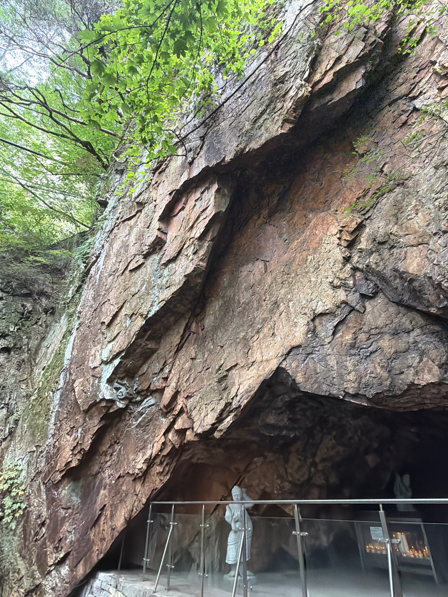 The image shows a view looking up at a rocky cliff face with lush green foliage overhanging from above. At the base of the cliff, there is an opening that appears to be a cave or grotto. A protective railing with glass panels is in place, leading towards the entrance of the grotto. Inside the grotto, there is a statue of a person, possibly religious in nature, and a rack of lit candles. The scene is set in a natural, outdoor environment, with the rock formations and vegetation suggesting a mountainous or forested area. The overall impression is one of a serene, spiritual, or historical site. The location is identified as the Unbo Lake Observation Deck in Dongducheon-si, South Korea.
