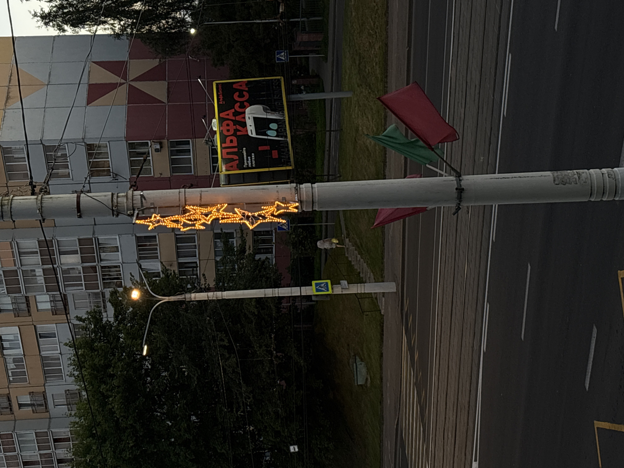 The image shows a street scene in Vitsebsk, Belarus. A tall residential building with a distinctive red and yellow geometric pattern on its facade dominates the background. In the middle ground, a lamppost is decorated with orange star-shaped lights. A billboard advertising "Alfa Kassa" is visible behind the lamppost. In the foreground, a pedestrian crossing with tram tracks is present. Three flags – one red, one green, and one red again – are attached to the lamppost. A person is seen walking up a set of stairs on the right side of the image. The sky is overcast.