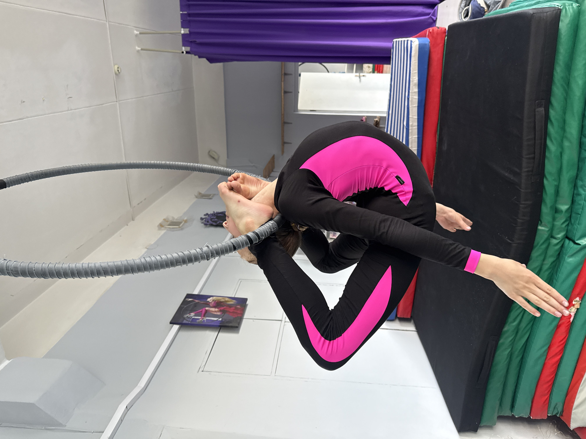 The image shows an aerialist performing a difficult pose on a suspended hoop in what appears to be an indoor training studio. The aerialist, wearing a black and neon pink leotard, is contorted into a highly flexible position, with their legs wrapped around the hoop and their body arched. Their feet are positioned above their head, and one hand is extended downwards, seemingly touching a stack of colorful crash mats below. The studio environment includes a white ceiling, grey walls, and a purple curtain in the background. A framed picture of an aerialist is visible on the wall. The crash mats are stacked in layers of black, green, and red, suggesting a gymnastics or aerial arts facility. The image does not provide specific details about the location within the city of Dnipro, Ukraine, beyond it being an indoor training space.