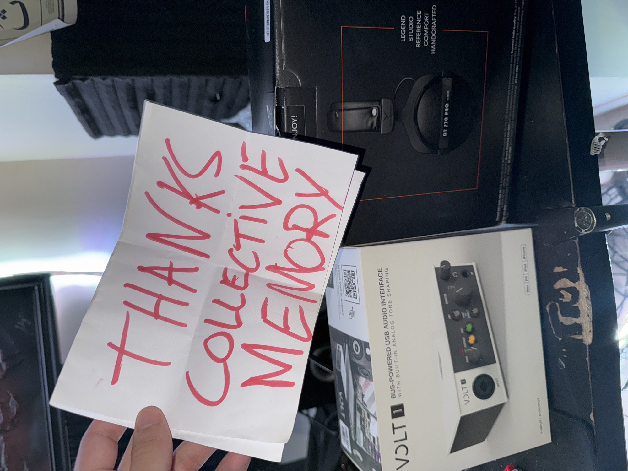 A person's hand holds a white paper note with the handwritten red text "THANKS COLLECTIVE MEMORY" prominently displayed in the foreground. The note is angled, partially covering two product boxes on a dark, possibly worn surface.
The upper box is black and features text and an image of a pair of black headphones. Visible text on this box includes "DT 770 PRO" and descriptors such as "LEGEND STUDIO REFERENCE COMFORT HANDCRAFTED". A word, possibly "ENJOY!", is partially visible near the top edge.
Below, a white and black box for an audio interface is visible. The model name "VOLT 1" is clearly printed. The box states it is a "BUS-POWERED USB AUDIO INTERFACE WITH BUILT-IN ANALOG TONE SHAPING" and details "1-INPUT / 2-OUTPUT". An image of the silver and black interface with its controls is shown, along with compatibility icons for "Mac PC iPad iPhone" and a QR code.
The scene depicts an indoor setting, likely a desk or workstation, where new audio equipment has been received. The handwritten note expresses gratitude.