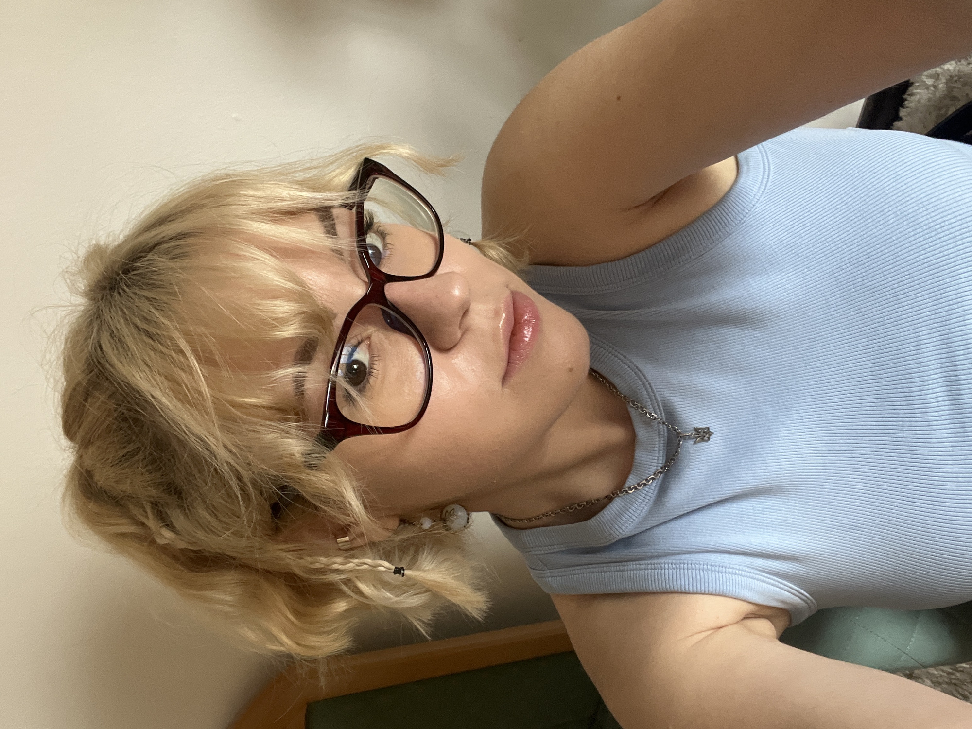 A young woman with blonde hair and glasses is posing for a selfie indoors. She is wearing a light blue ribbed tank top and a silver necklace with a pendant. Her hair is styled with loose waves and small braids on the side. She is looking directly at the camera with a slight smirk. The background is a plain white wall. The location is not provided in the image.
